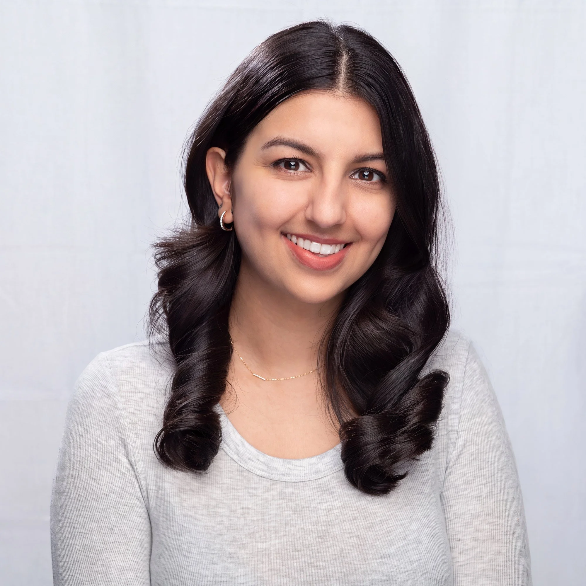 Professional headshot of a young South Asian woman in a light-colored sweater against a natural muslin cloth backdrop.