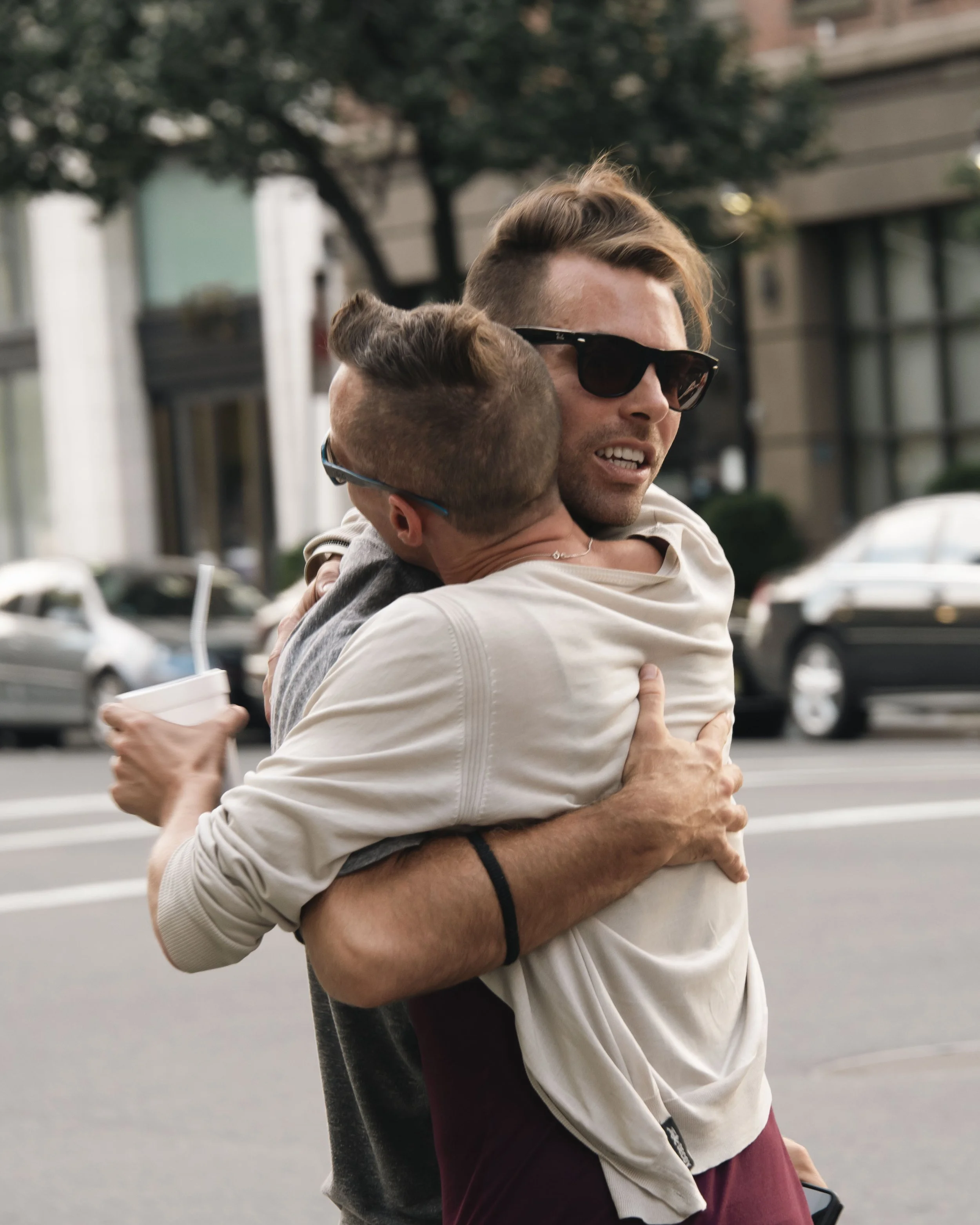 Two young men hugging on a city street, wearing sunglasses, with cars and buildings in the background.