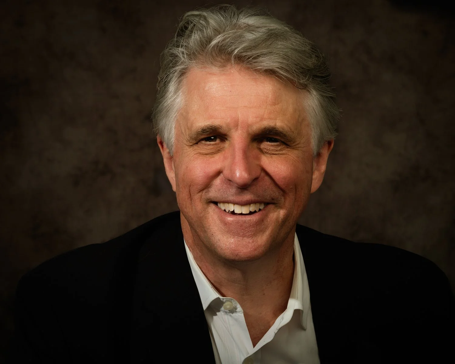 A smiling middle-aged man with gray hair, wearing a black blazer and white collared shirt, against a dark background.