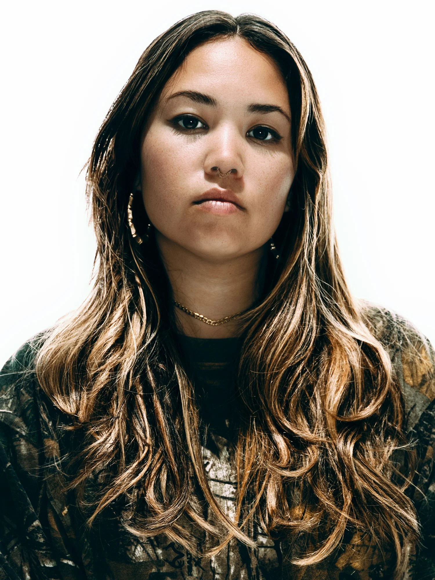 Portrait of young, expressionless woman with long, light brown hair, earrings, neck chain, and nose piercing, wearing a camo-pattern jacket. Photographed with hard, directional studio light on a white background.