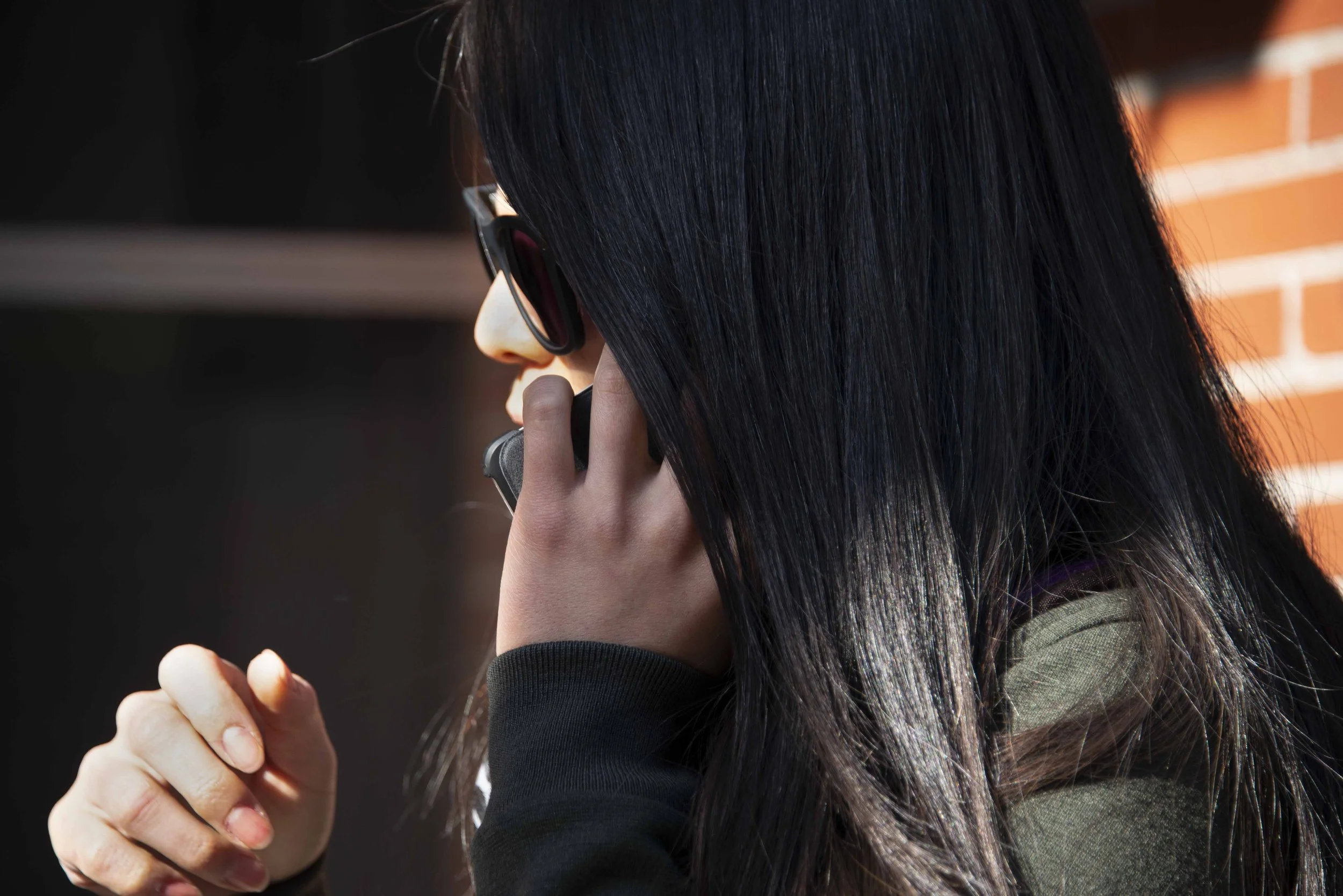 A woman with long black hair and sunglasses is talking on a cellphone and holding a small object in her hand.