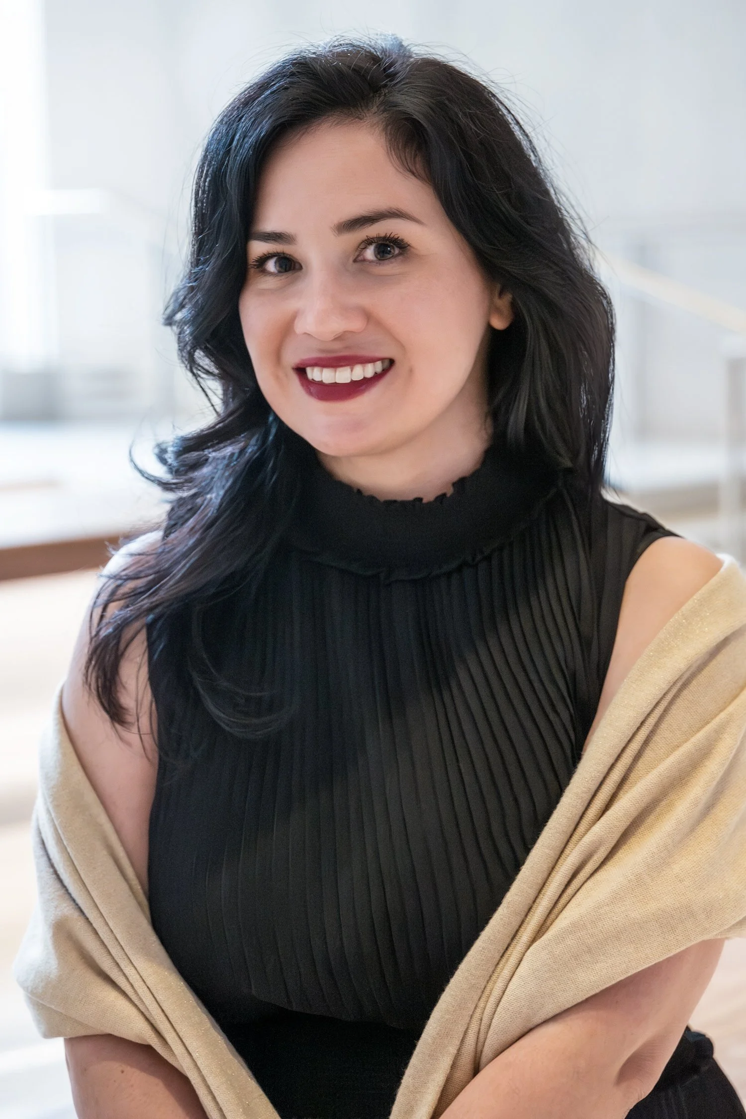 Portrait of young woman with long, dark hair and red lips wearing a light tan shawl over a black high-neck top. She’s seated next to a window with shallow focus blurring the background.