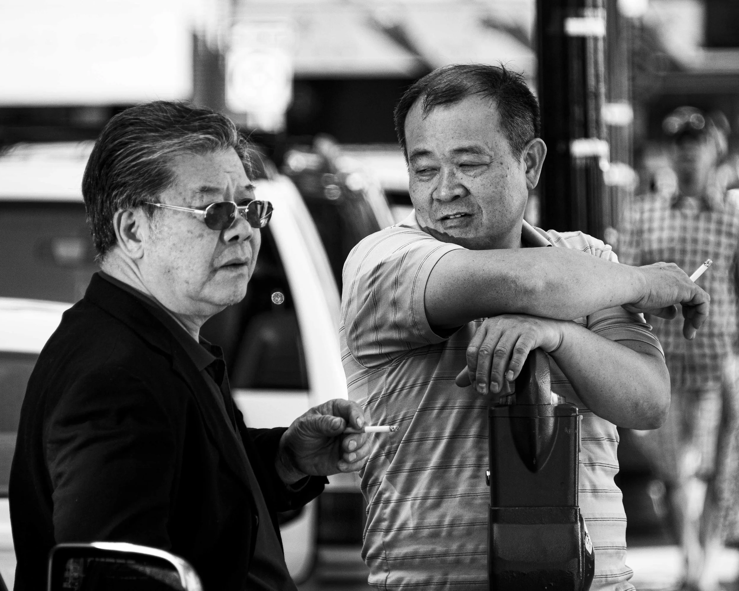 Two men, one wearing sunglasses and the other not, are sitting at an outdoor table, engaged in conversation, with a street scene in the background.