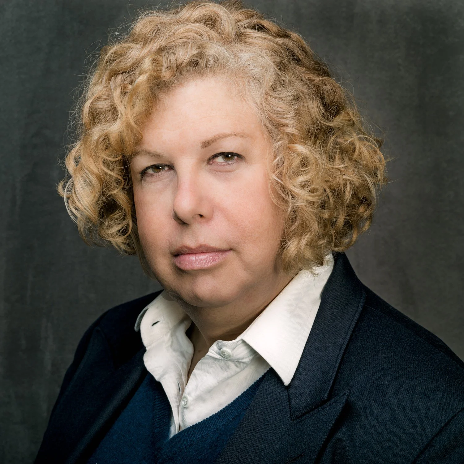Portrait of a trans woman with curly, blonde hair, wearing a dark blazer with a blue v0neck sweater over a white, collared shirt against a drk gray background.
