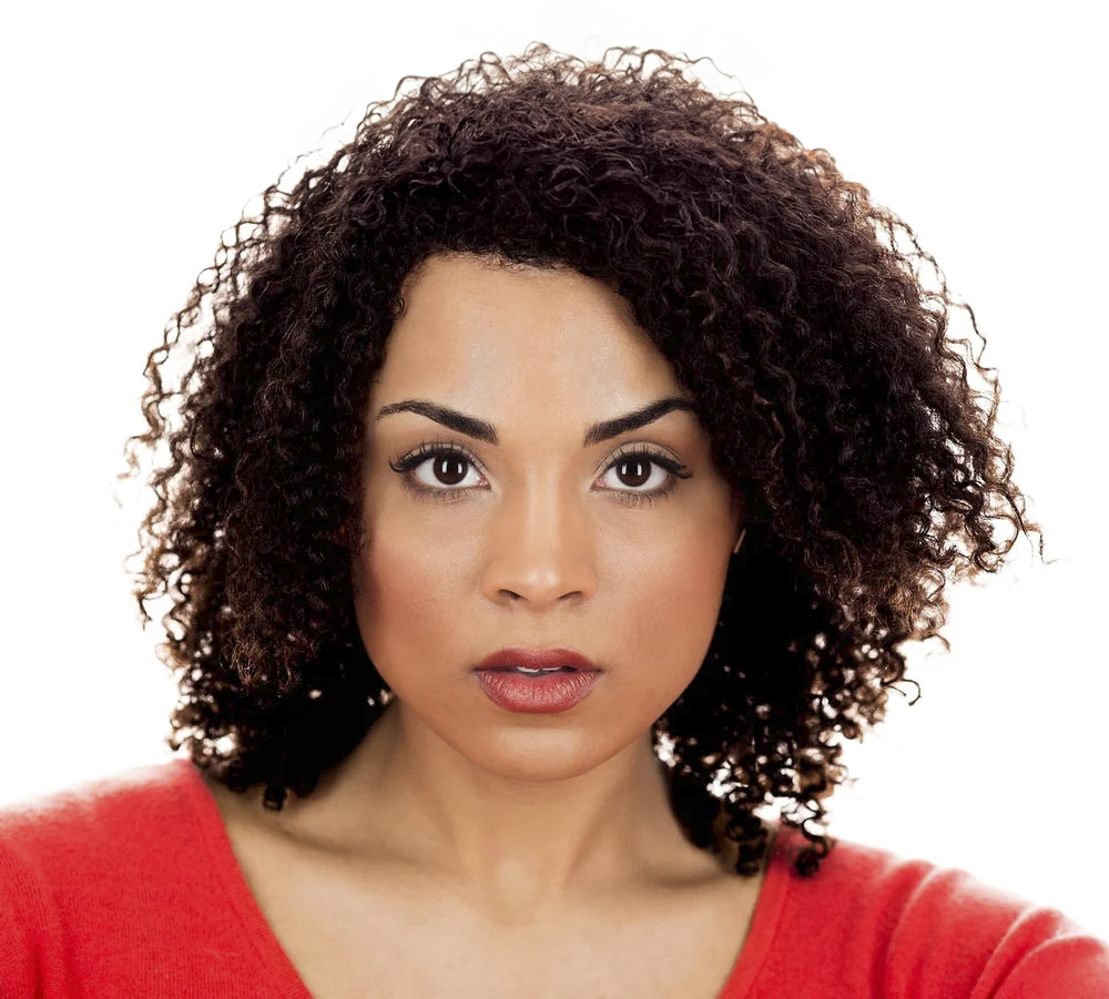 A Black woman with dark curly hair, wearing a red top, looking directly into the camera with a neutral expression against a white background.