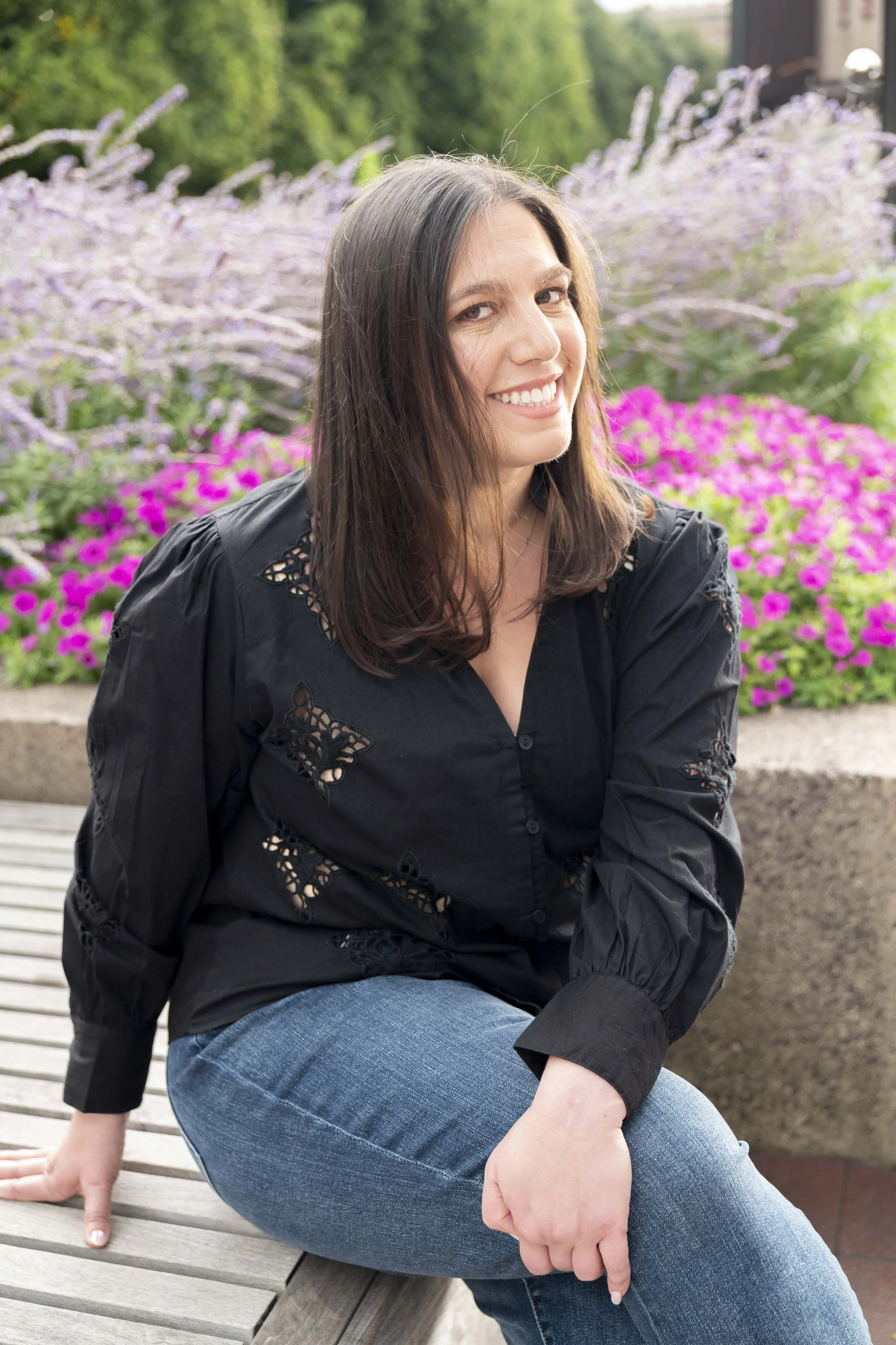 A woman with brown hair smiling, sitting on a wooden bench outdoors with pink flowers and green bushes in the background.