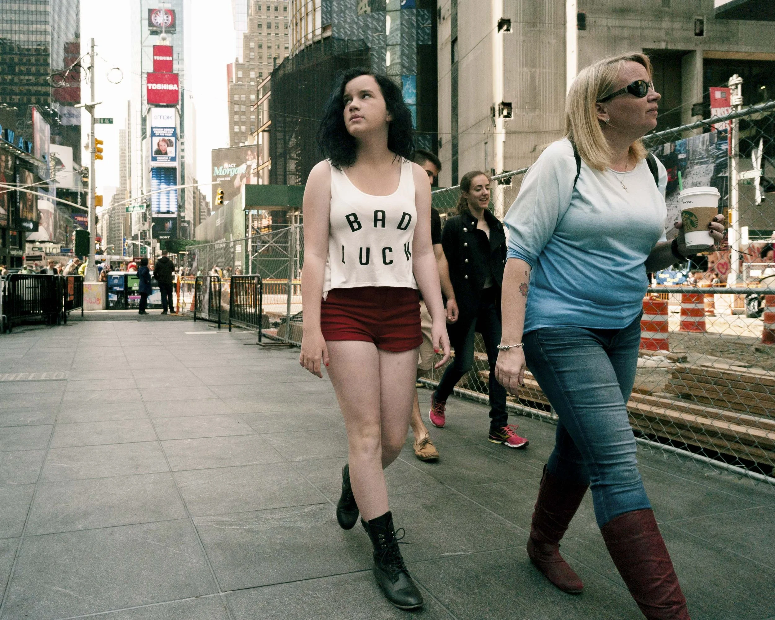 A group of people walking on a city street during the daytime, with tall buildings and billboards in the background.