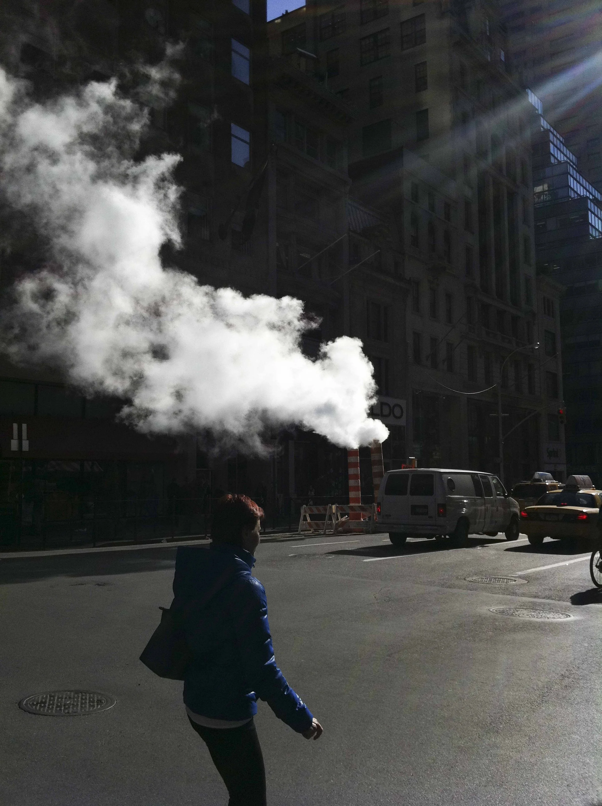 A person walking on a city street with steam rising from underground vents, with tall buildings and cars visible in the background.