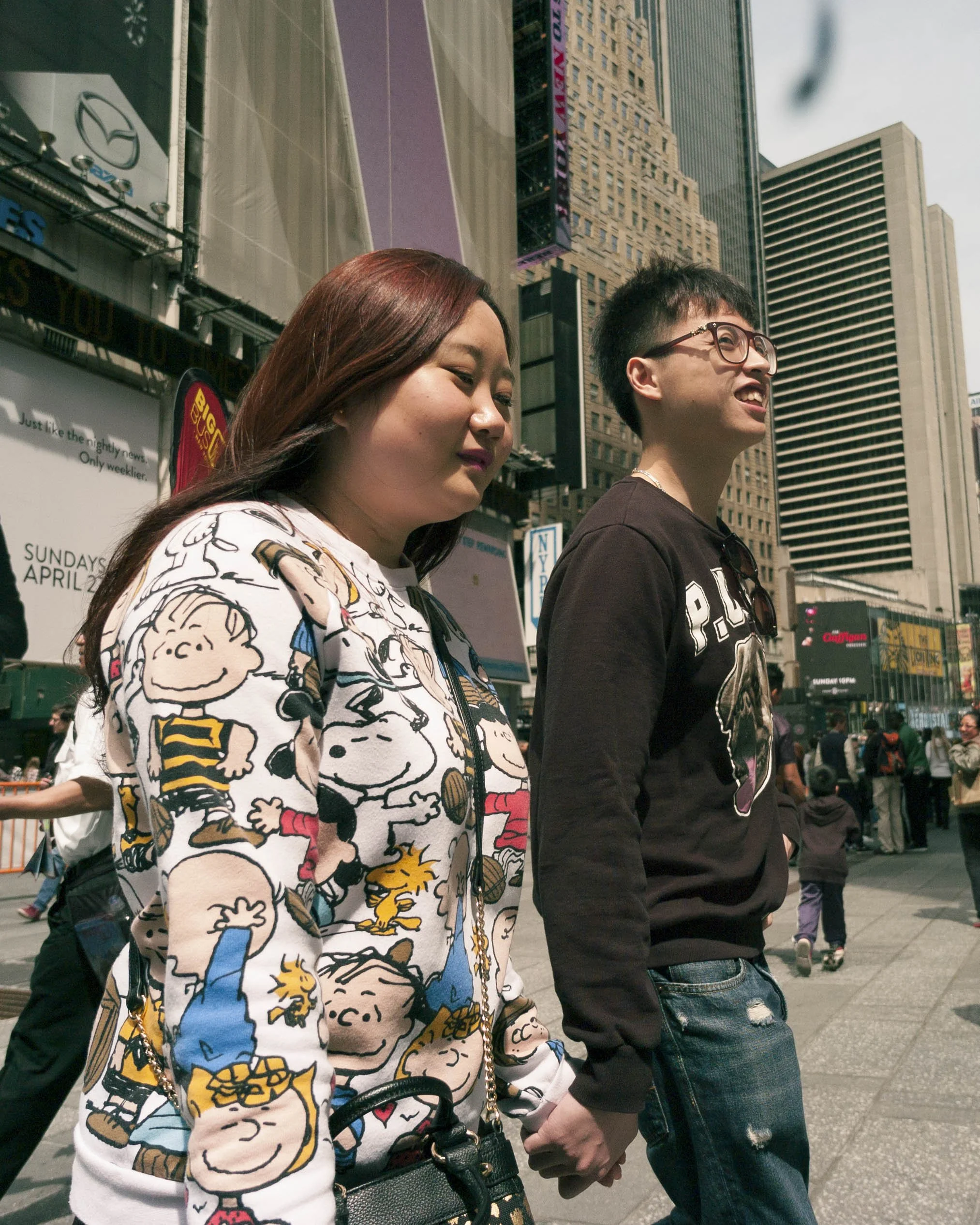 Two young people holding hands walk in a busy city street, tall buildings and digital billboards in the background.