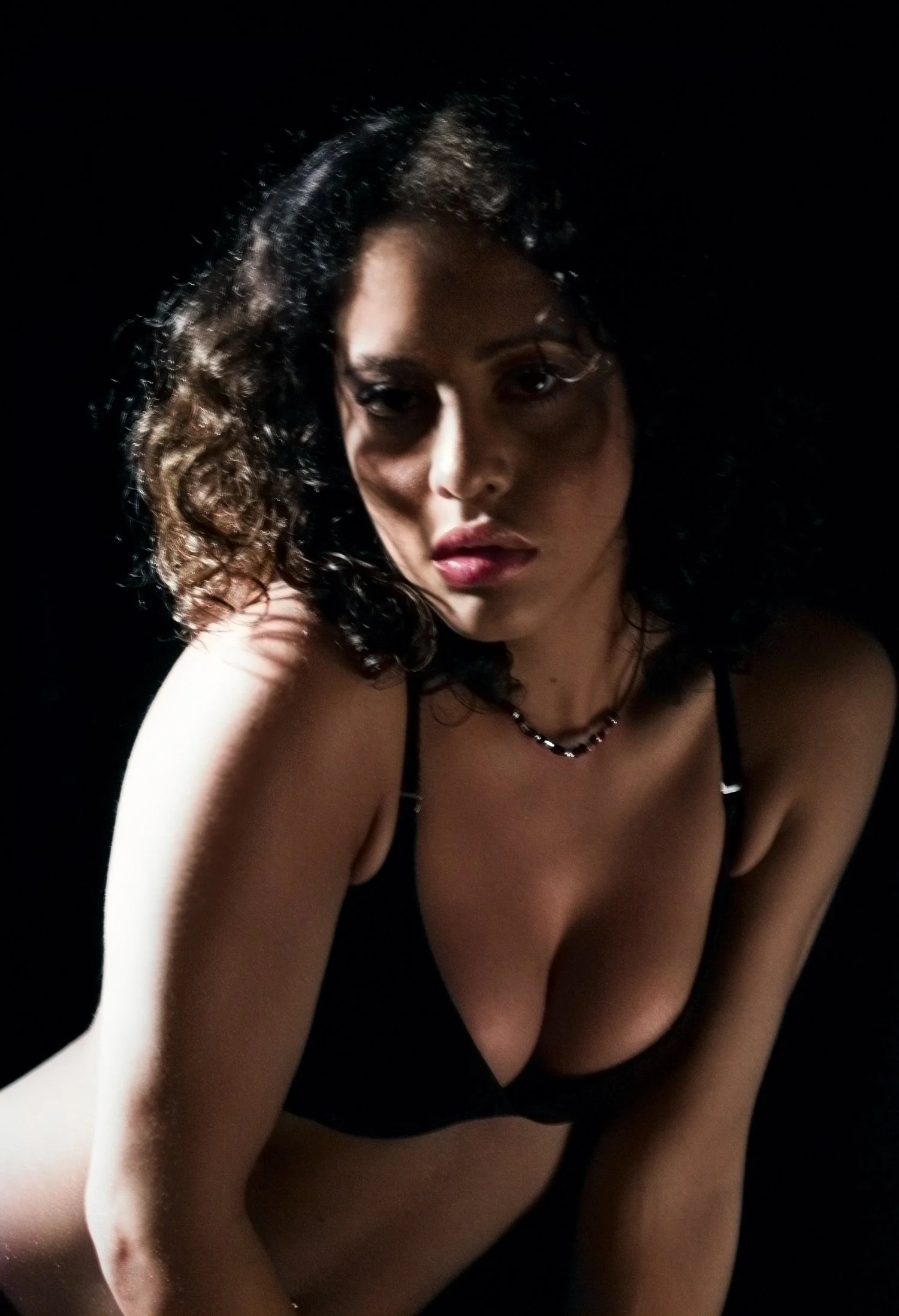 Close-up portrait of a woman with curly black hair, wearing a black top and a beaded necklace, against a dark background.