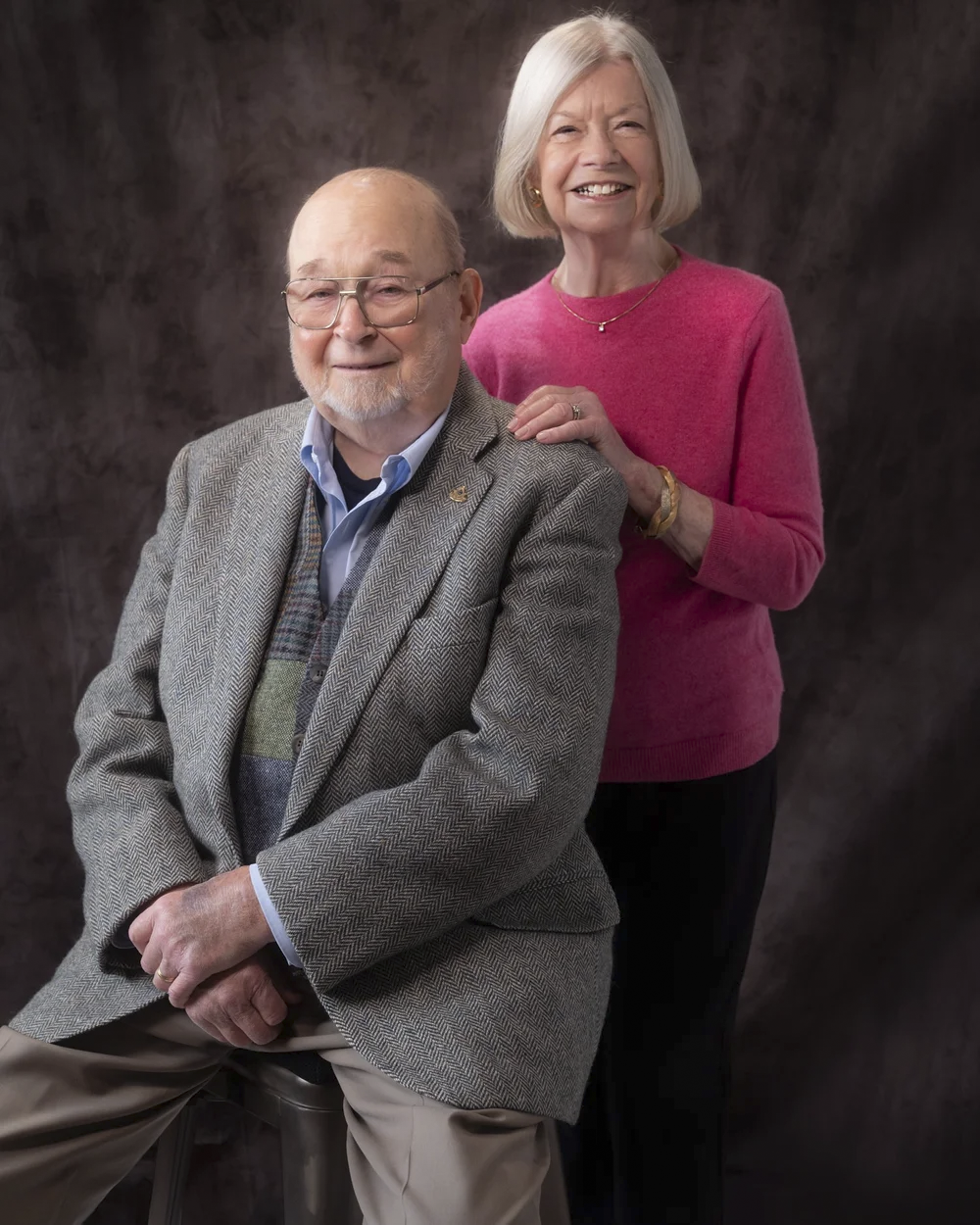 An elderly man with glasses and a woman with white hair pose together against a dark, textured background. The man is seated, wearing a gray herringbone blazer, blue shirt, and beige pants. The woman stands behind him, resting her hand on his shoulde
