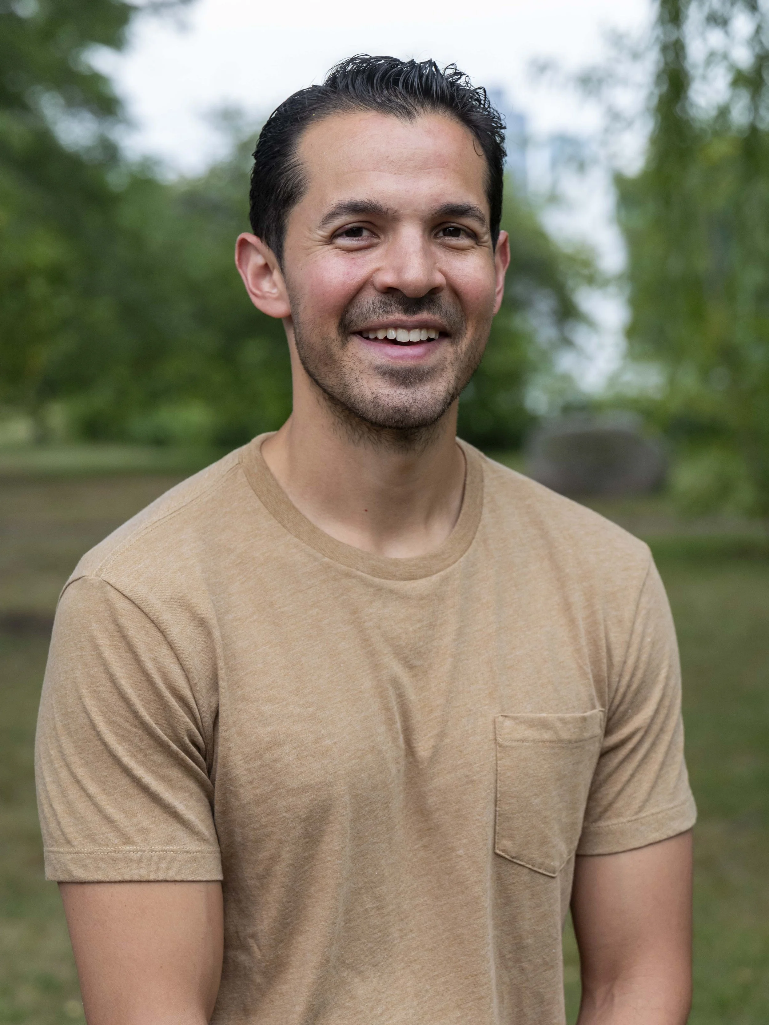 A smiling man with dark, slicked-back hair, wearing a beige t-shirt, standing outdoors in a park with green trees in the background.
