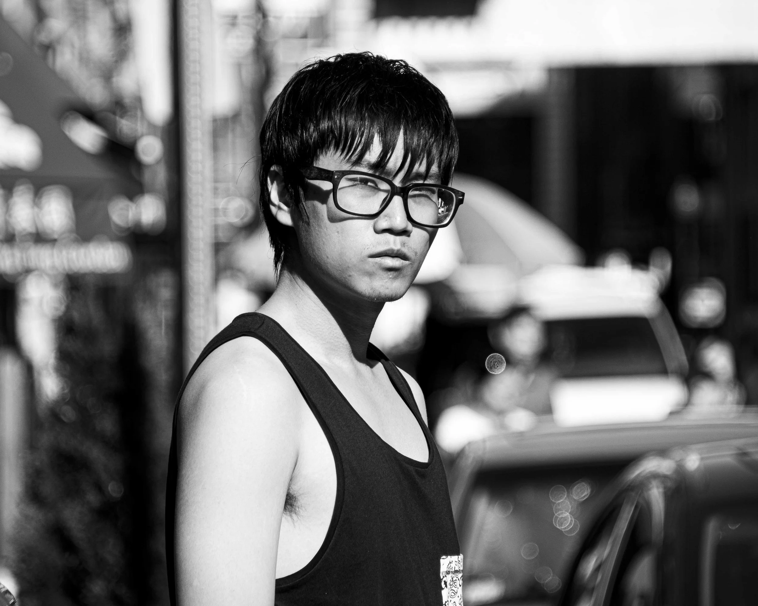 A person with short, dark hair and glasses looking over their shoulder in a black and white photo, with a blurred urban background.