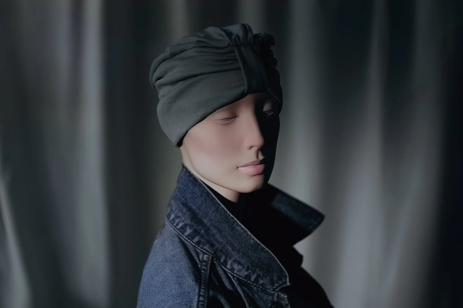 A woman with fair skin is wearing a gray head wrap and a dark denim jacket, with her eyes closed and her face turned slightly to the side against a blurred background.
