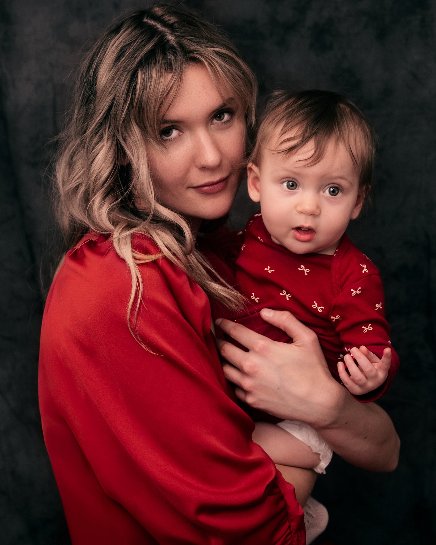 A young mother with her infant daughter, both dressed in red outfits, photographed in classic studio portrait style for Mother’s Day.