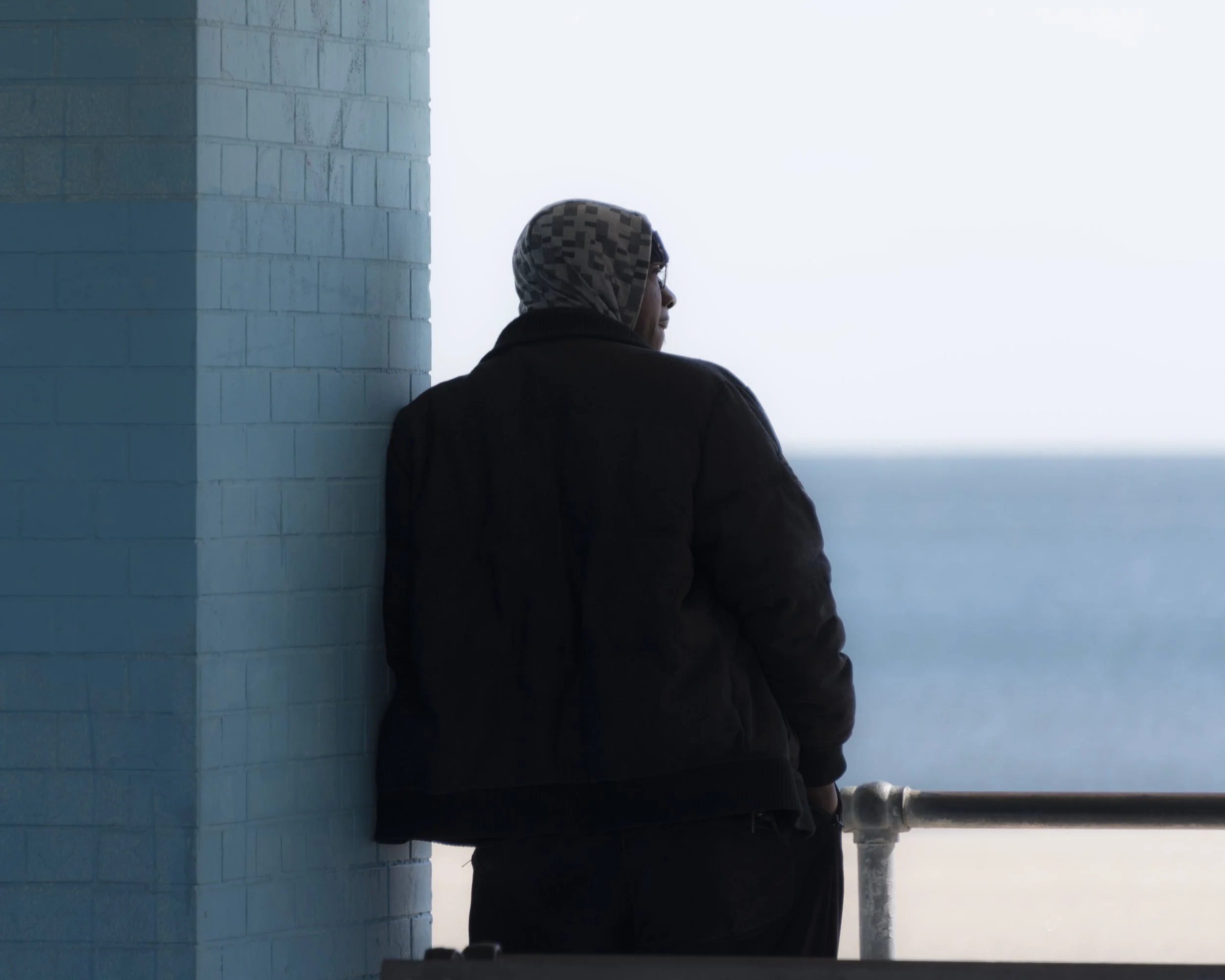 A person with a headscarf standing next to a light blue brick wall, looking towards the ocean or a large body of water in the distance.
