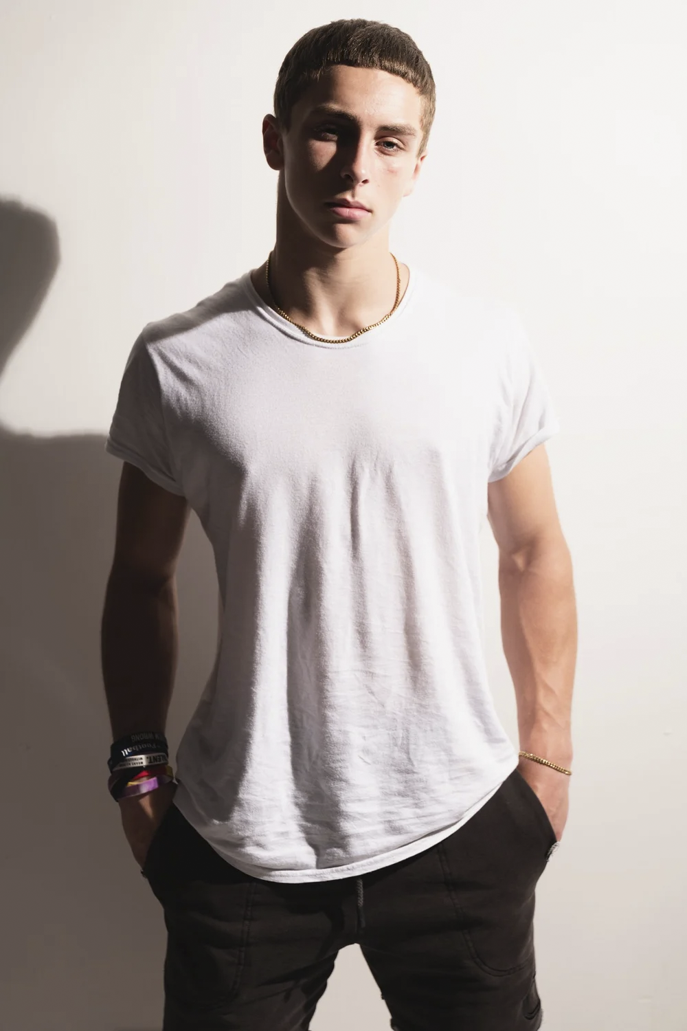 Portrait of a young male athlete in a loose-fitting white T-shirt, hands in pocket, seen in dramatic directional light.