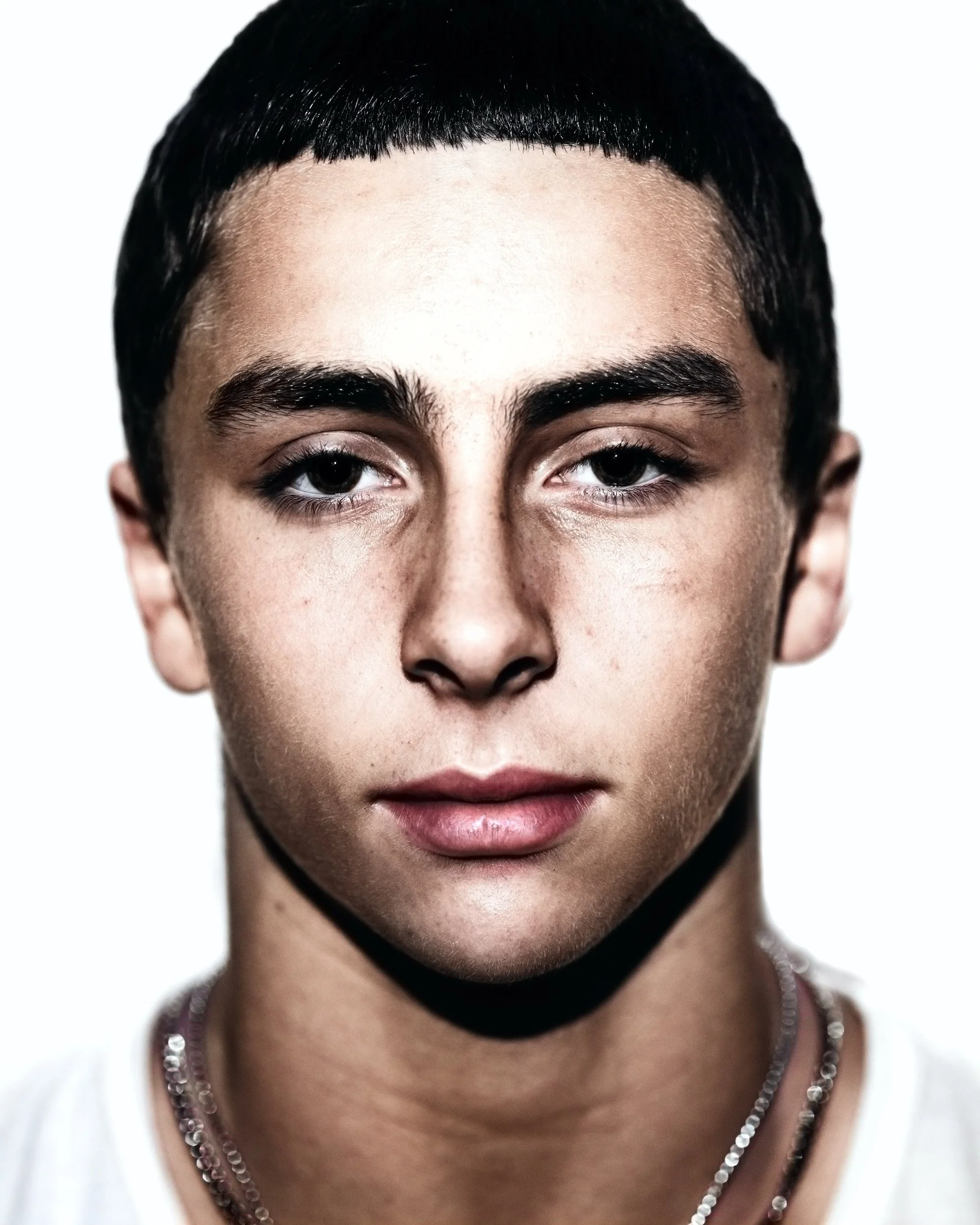 Close-up portrait of a person with short, dark hair, thick eyebrows, and brown eyes, wearing layered silver necklaces.