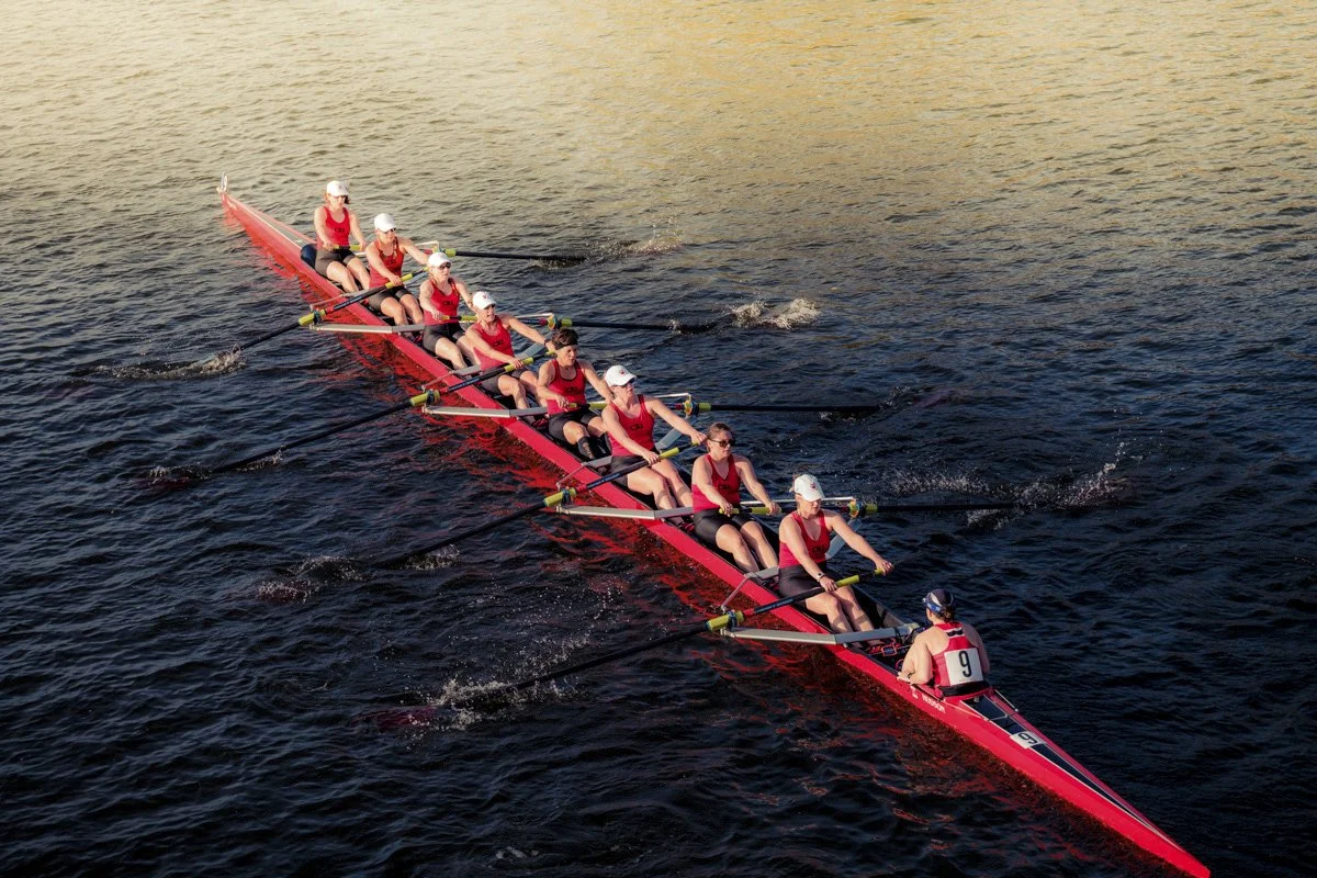 A row of nine women in red athletic shirts and white caps, with a coxswain at the front, rowing a red boat on dark water during sunset.