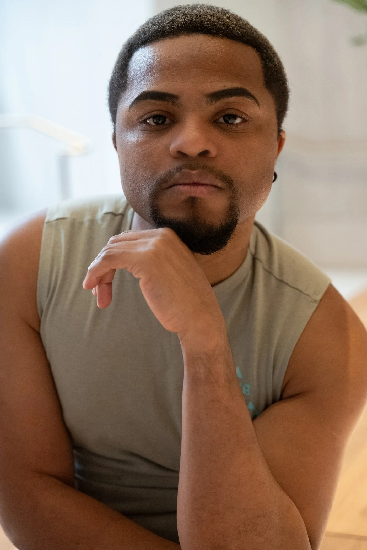 A young man with dark hair, beard, and mustache, wearing a sleeveless shirt, resting his chin on his hand and looking into the camera.