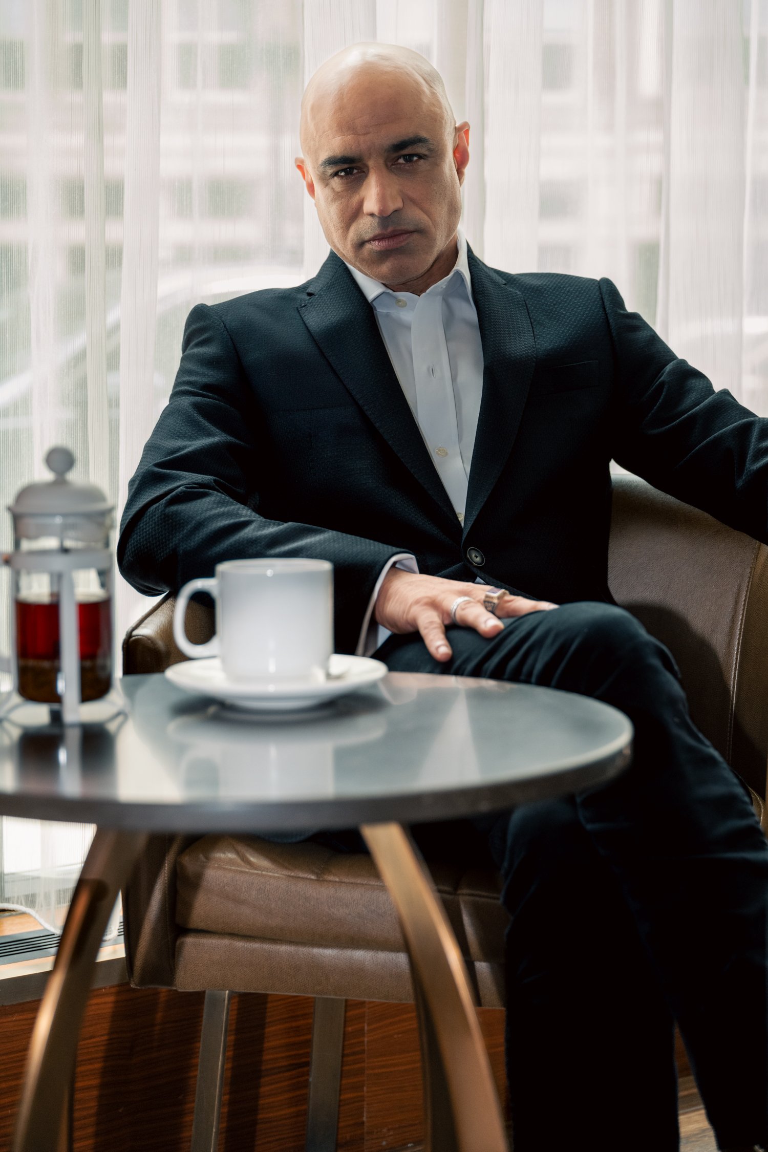 A bald man in a dark suit and white shirt sitting at a table, looking serious. There are coffee mugs and a juice bottle on the table, with sheer curtains and a large window in the background.