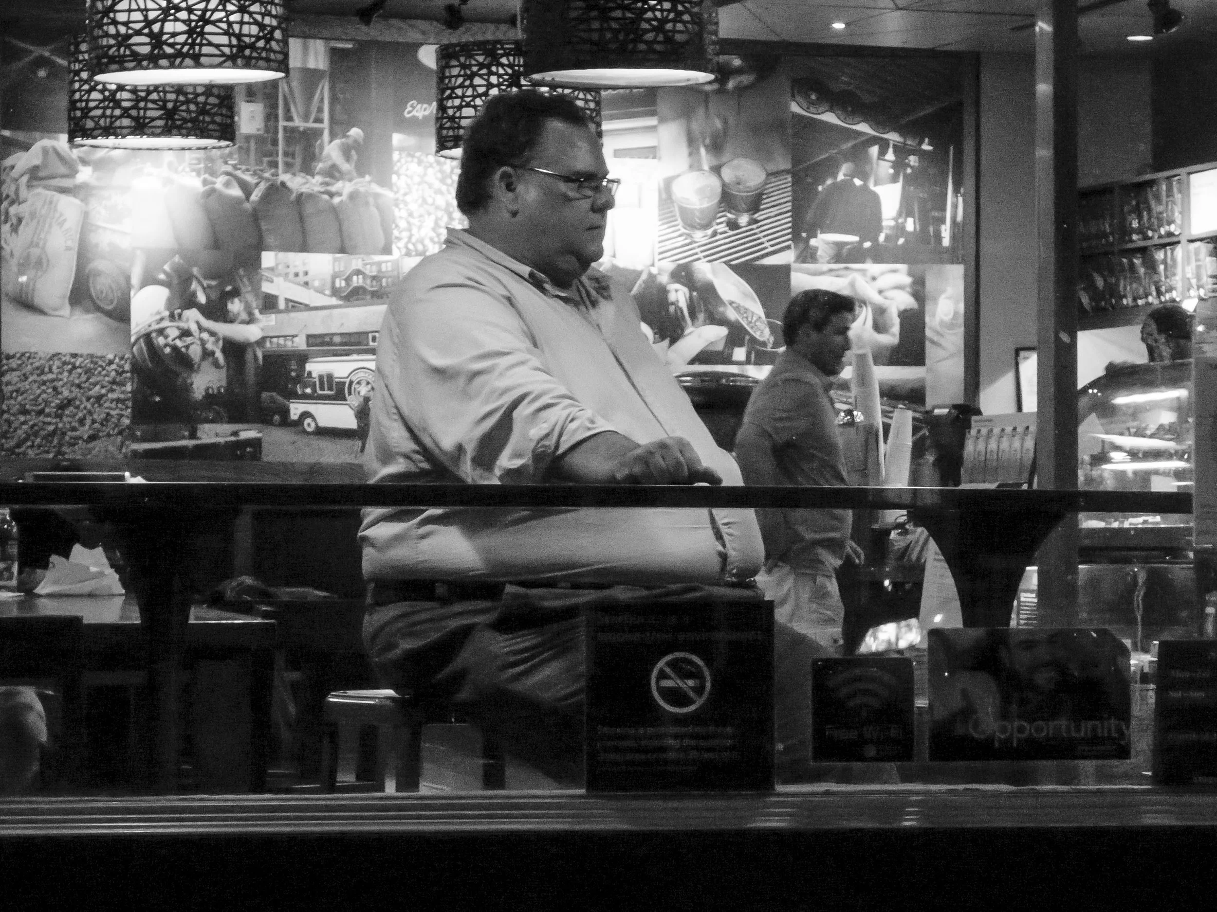 A black and white photo of two men sitting at a bar, with a busy wall decorated with various photographs and images in the background. The man in the foreground is wearing glasses and a collared shirt, and the man in the background is reading somethi