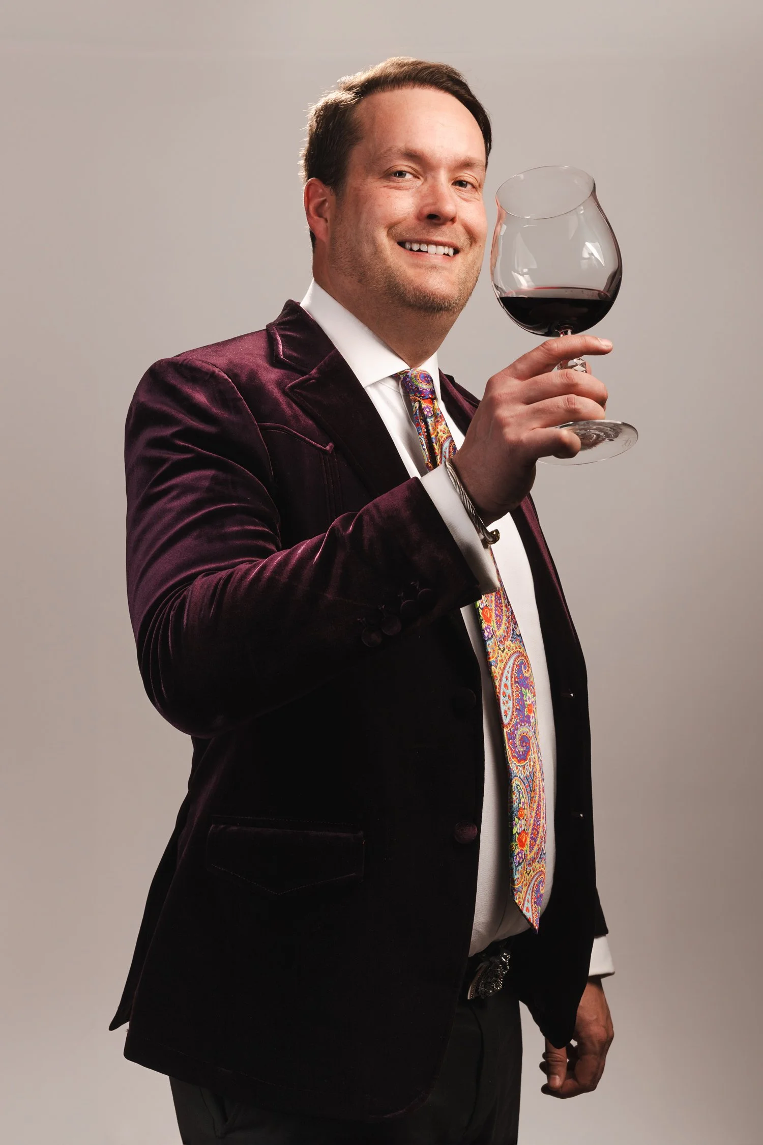 A man in a dark purple velvet blazer, white shirt, and colorful paisley tie holding a glass of red wine, smiling at the camera.