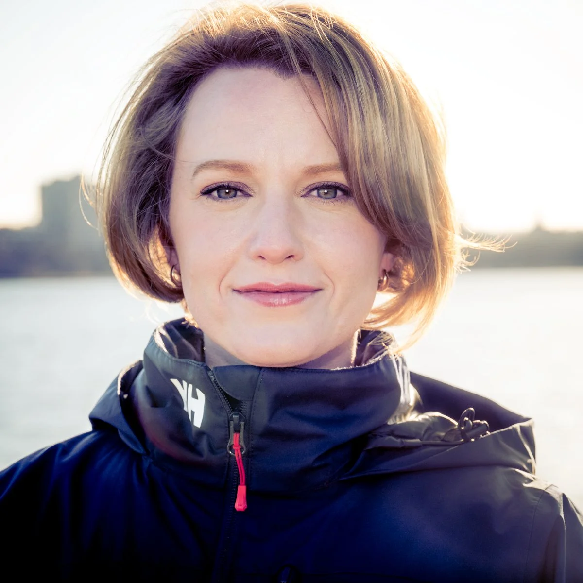 Close-up of a woman with short light brown hair, wearing a black outdoor jacket, outdoors near a body of water, with a city skyline in the background.
