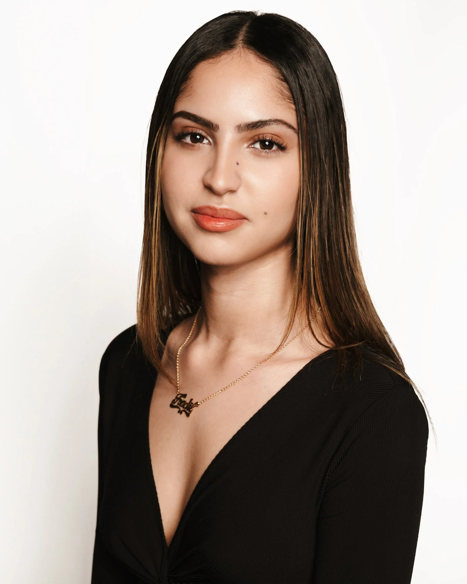 Portrait of a young woman of North African descent with long, straight, brown hair wearing an open-neck black top with a gold chain and pendant. She wears a subtle smile as she looks intently into the camera. The background is pure white.