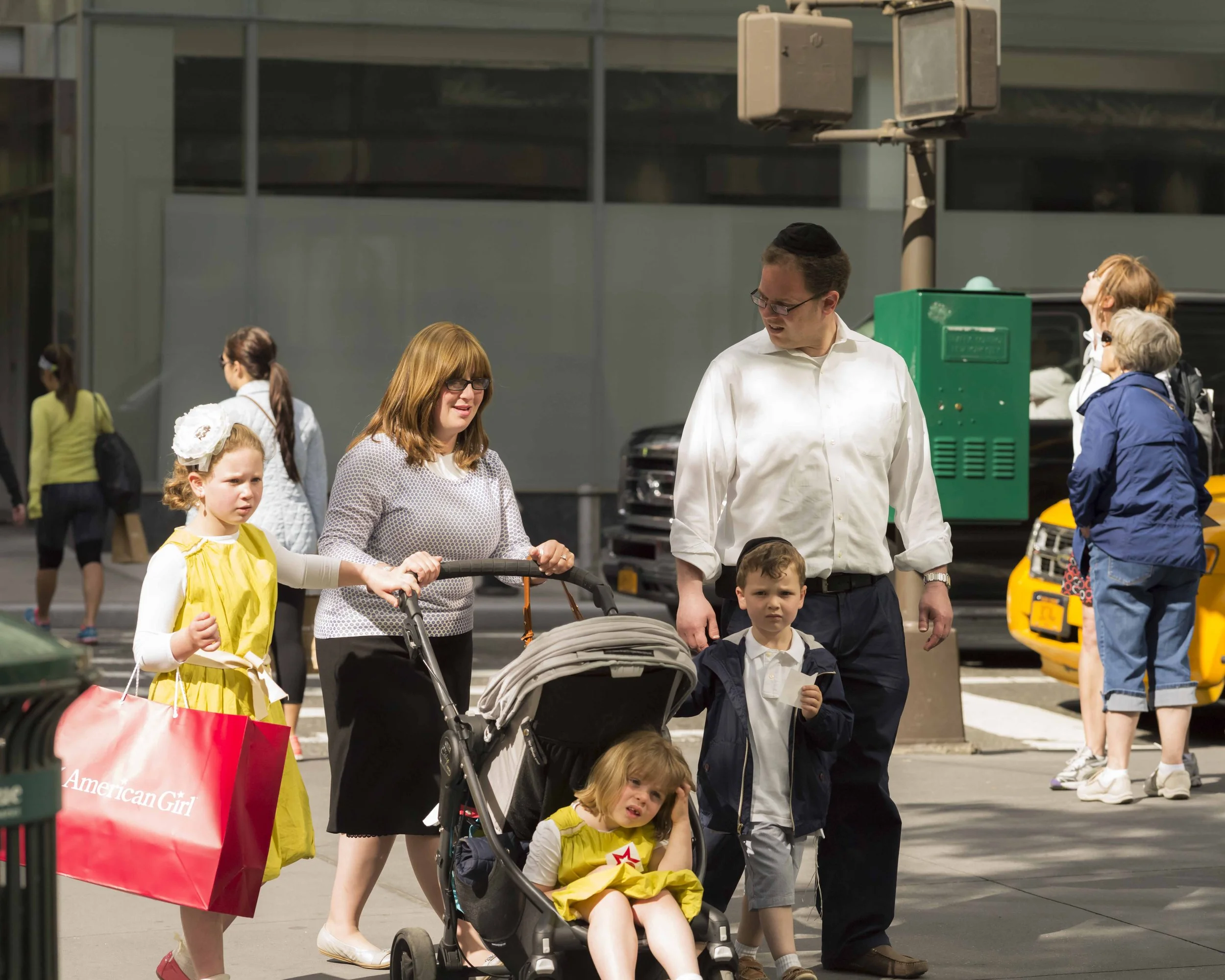 A group of people crossing the street, including a woman with a stroller, children, and adults, in front of a building and parked cars.