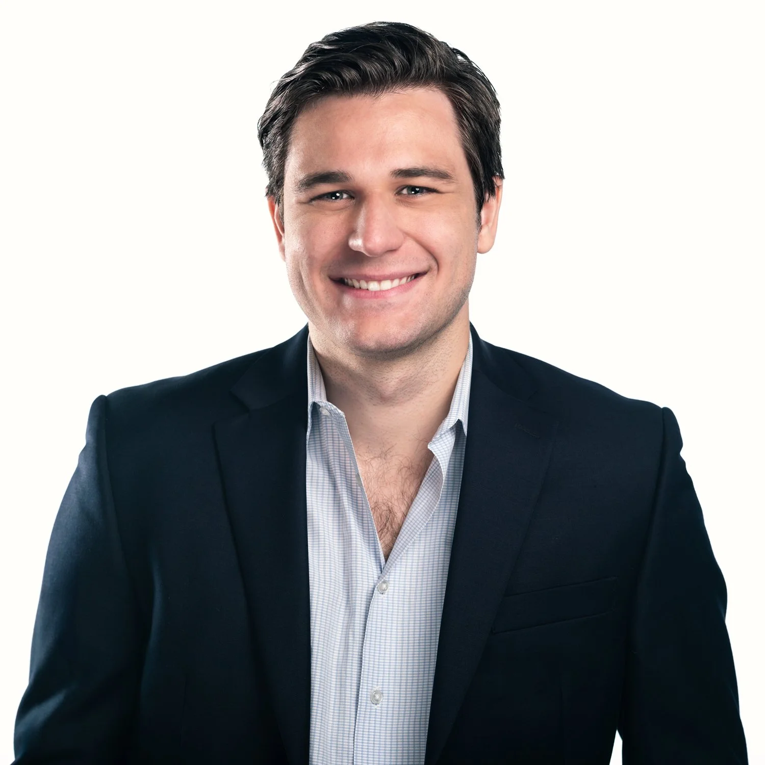 Headshot of a smiling, young male executive, wearing an open-collar white shirt and a blue suit jacket. It is shot on a white background in a studio setting.