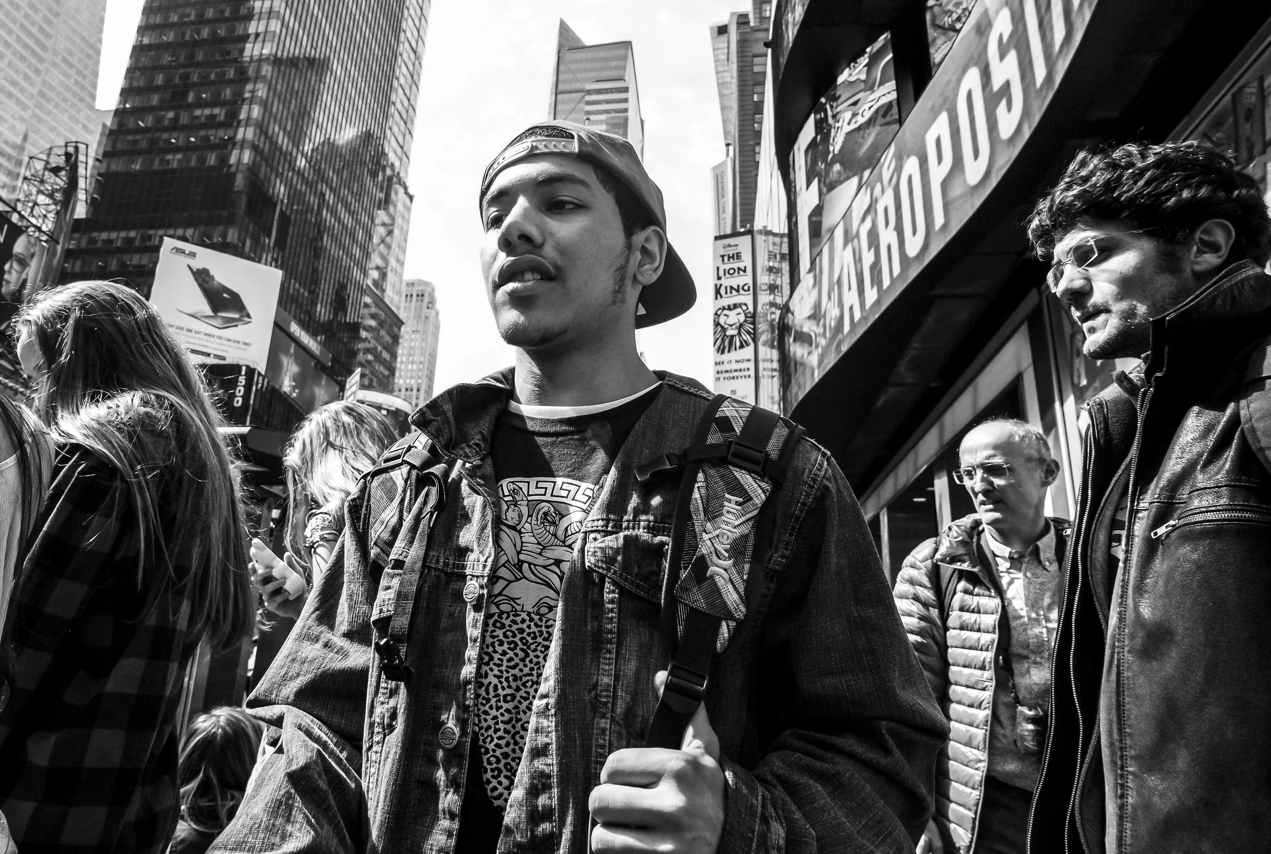 Black and white photo of a young man in a denim jacket and cap standing in Times Square, New York City, surrounded by other pedestrians and tall buildings with billboards and signs.