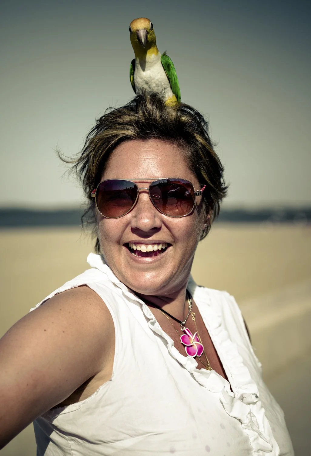 A woman with short hair wearing sunglasses, smiling, and standing on a beach with a parrot perched on her head.