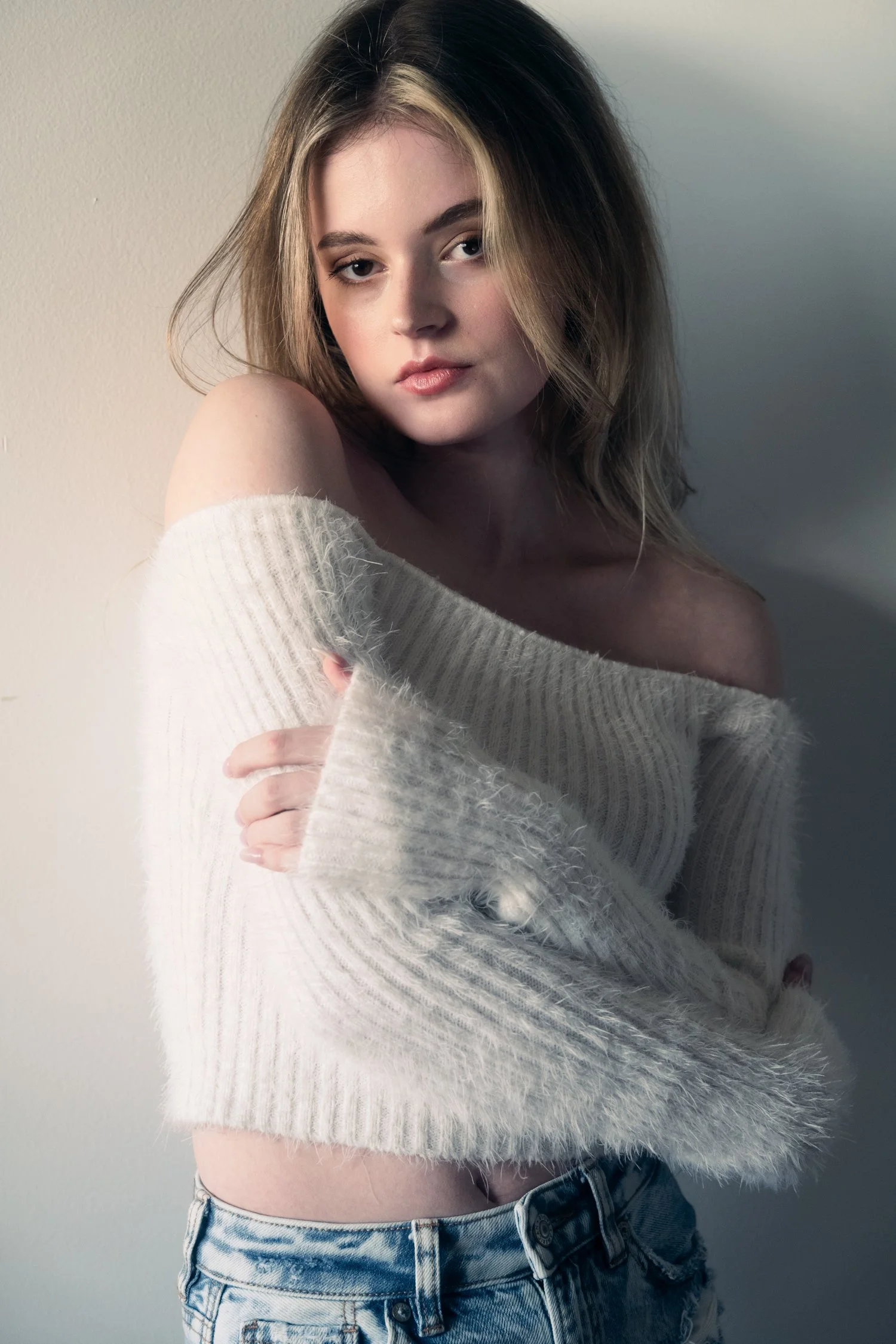 Young woman poses for her model card digitals in a white, off-the-shoulder sweater with blue denim in a studio setting featuring dynamic shadows.