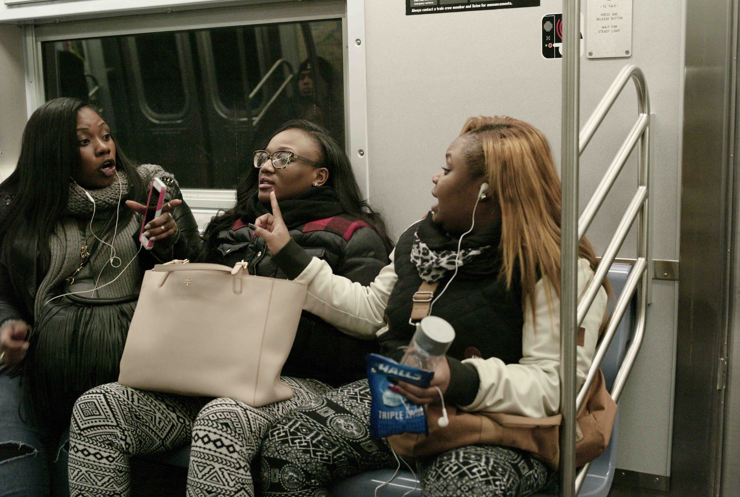 Three women sitting on a subway train, engaged in conversation. One woman has long black hair, another has curly hair and glasses, and the third has reddish-blonde hair. They are all wearing earphones and have bags with them.