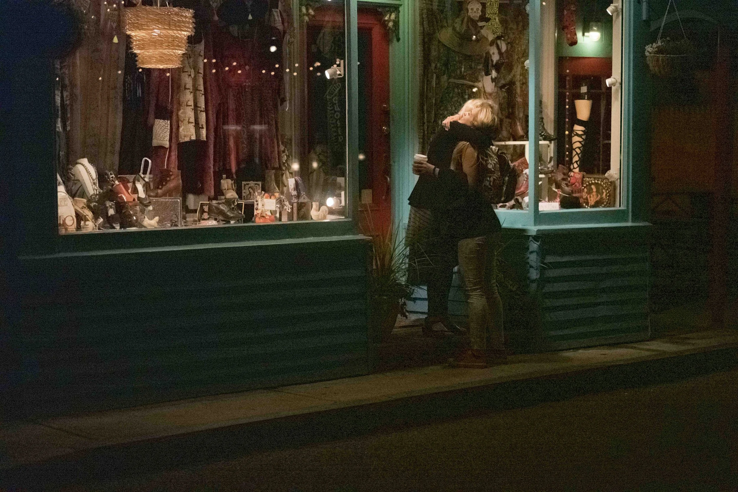 Nighttime view of a storefront window display with clothing and accessories, with two women embracing outside.