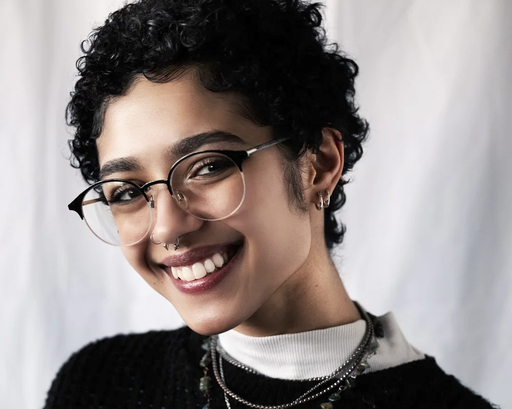 Close-up portrait of a young woman with short curly black hair, wearing glasses, earrings, a septum piercing, layered necklaces, a white turtleneck, and a black sweater, smiling against a plain white background.