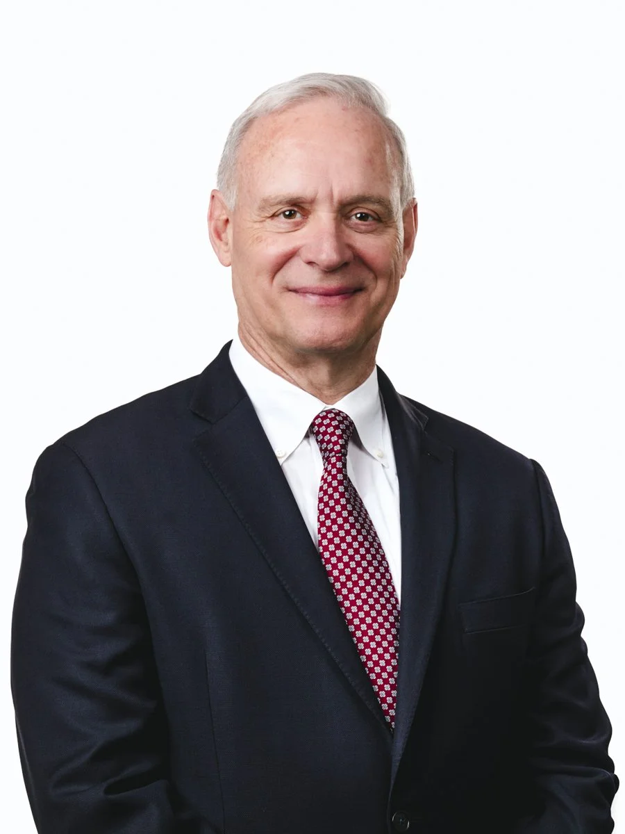 Executive headshot featuring an older gentleman with short gray hair, wearing a dark suit with a white shirt and red tie, shot against a white backdrop.