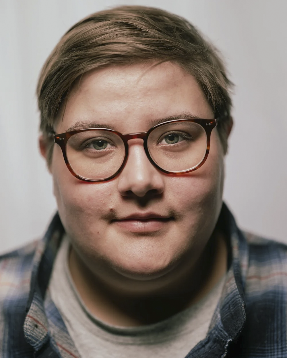 Close-up of a person with short brown hair and glasses, wearing a plaid shirt, looking directly at the camera.