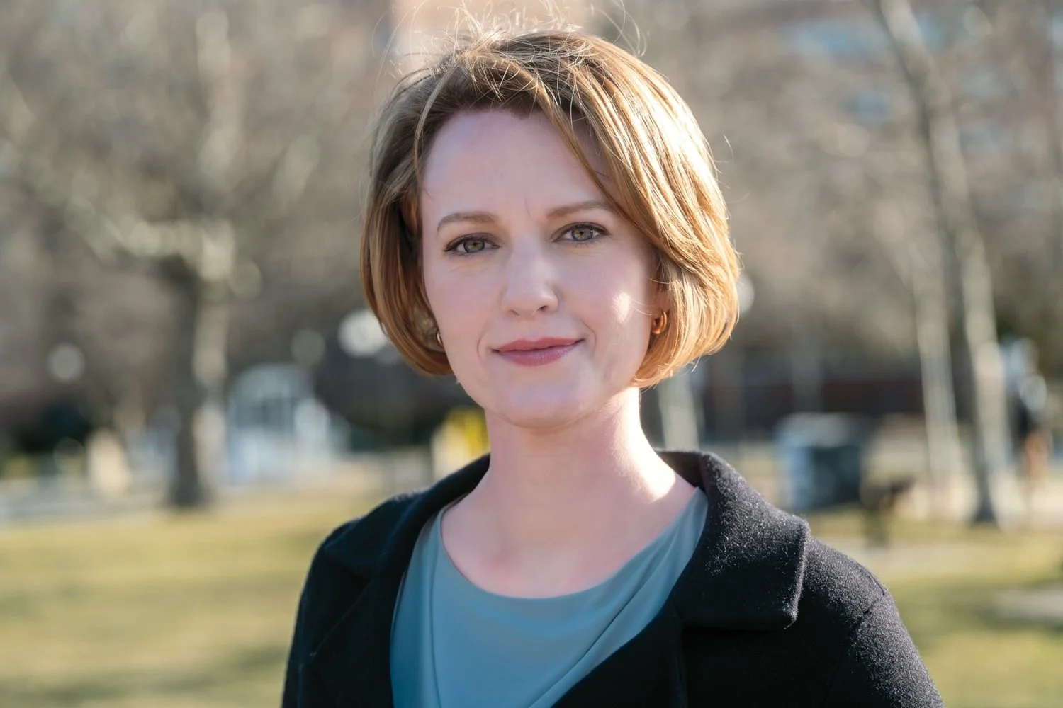 A woman with short auburn hair wearing a light blue top and black coat outdoors on a sunny day.