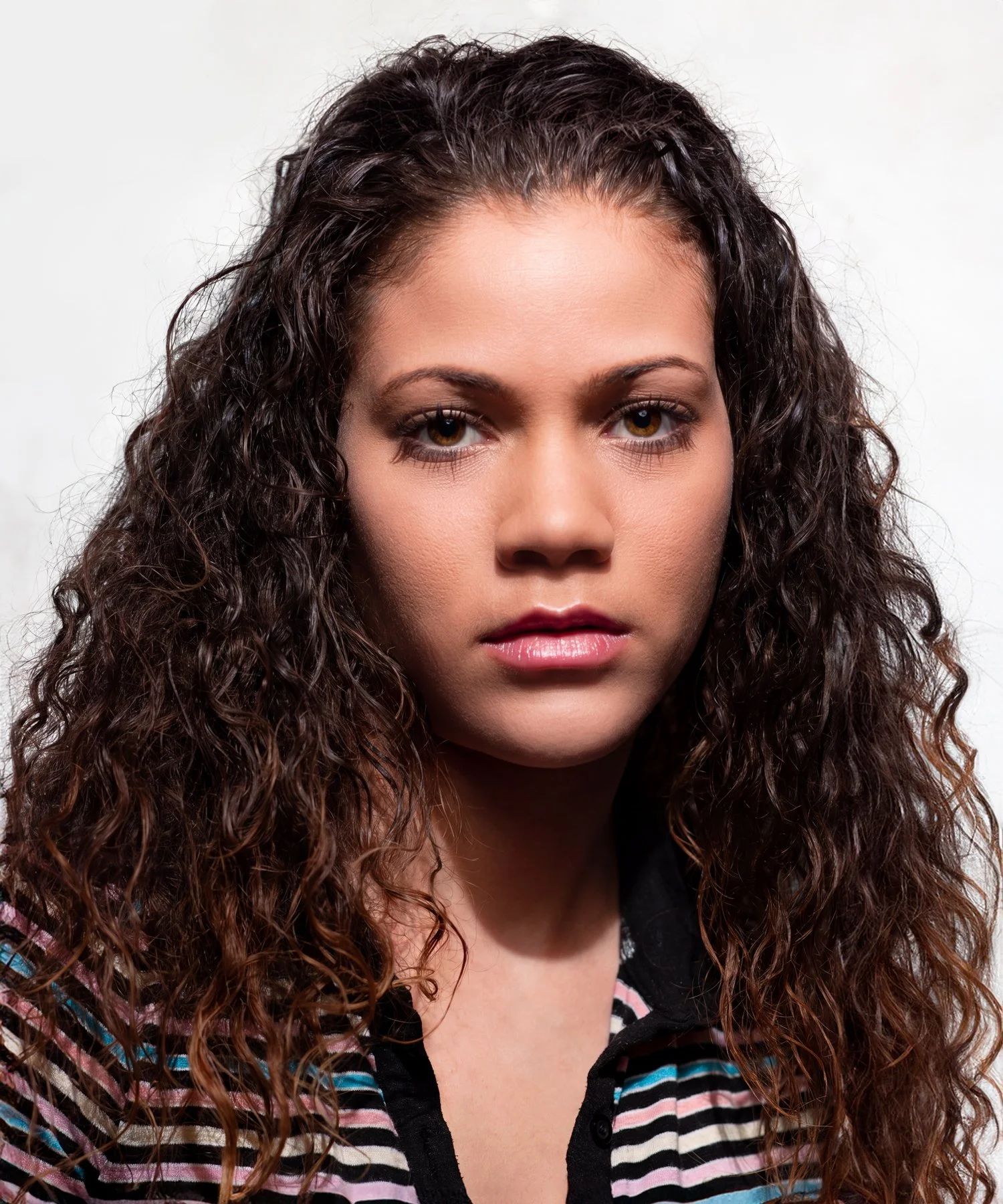 Studio portrait of young Latina female with long, curly hair. and blue, pink, white, and black striped shirt. Shot with beauty light against a white background.