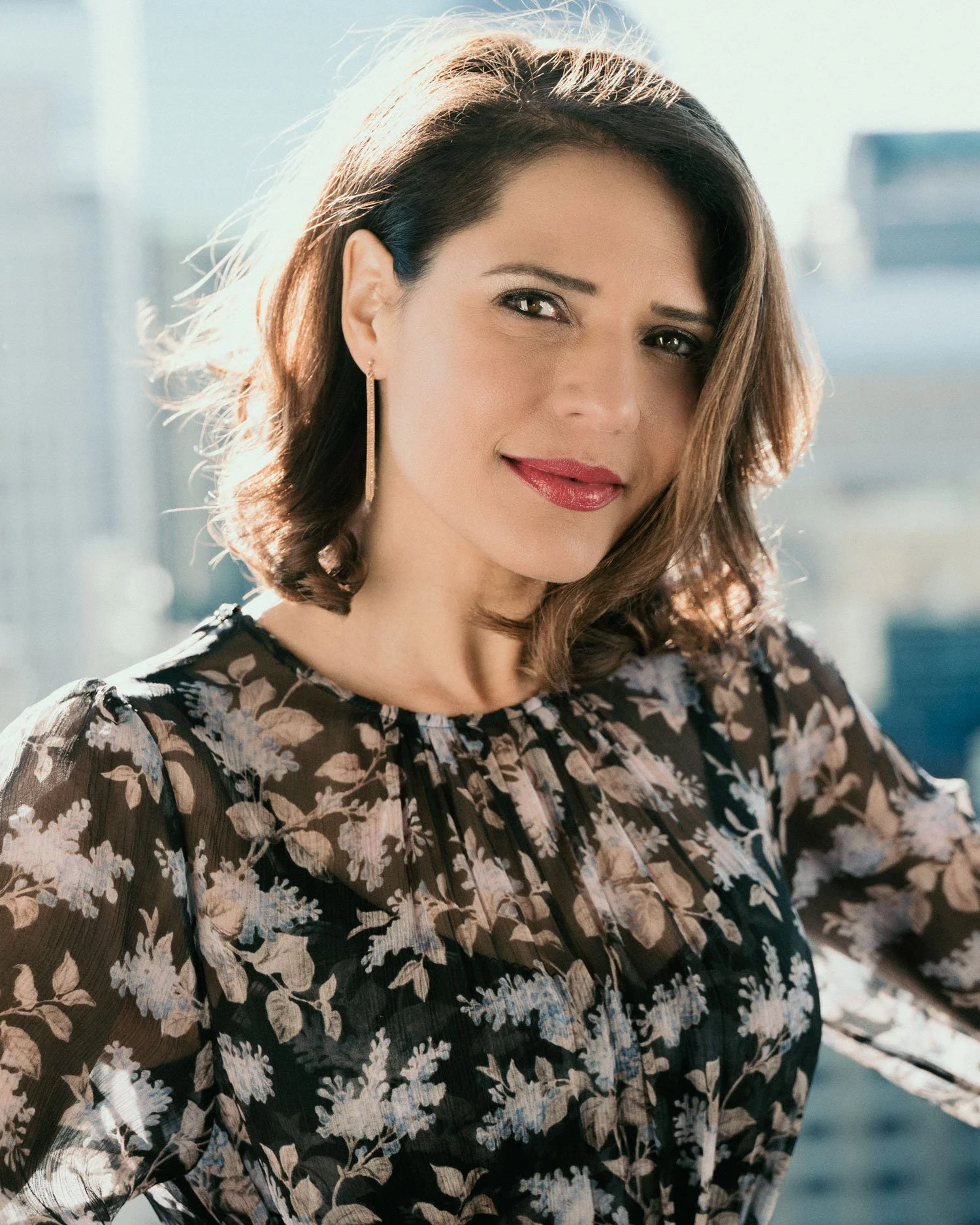 Actress Monique Gabriella Curnen in a natural light portrait shot outdoors in sunlight against a city background. She has shoulder-length brown hair and red lips and wear a dress with sheer fabric in brown and white floral print.