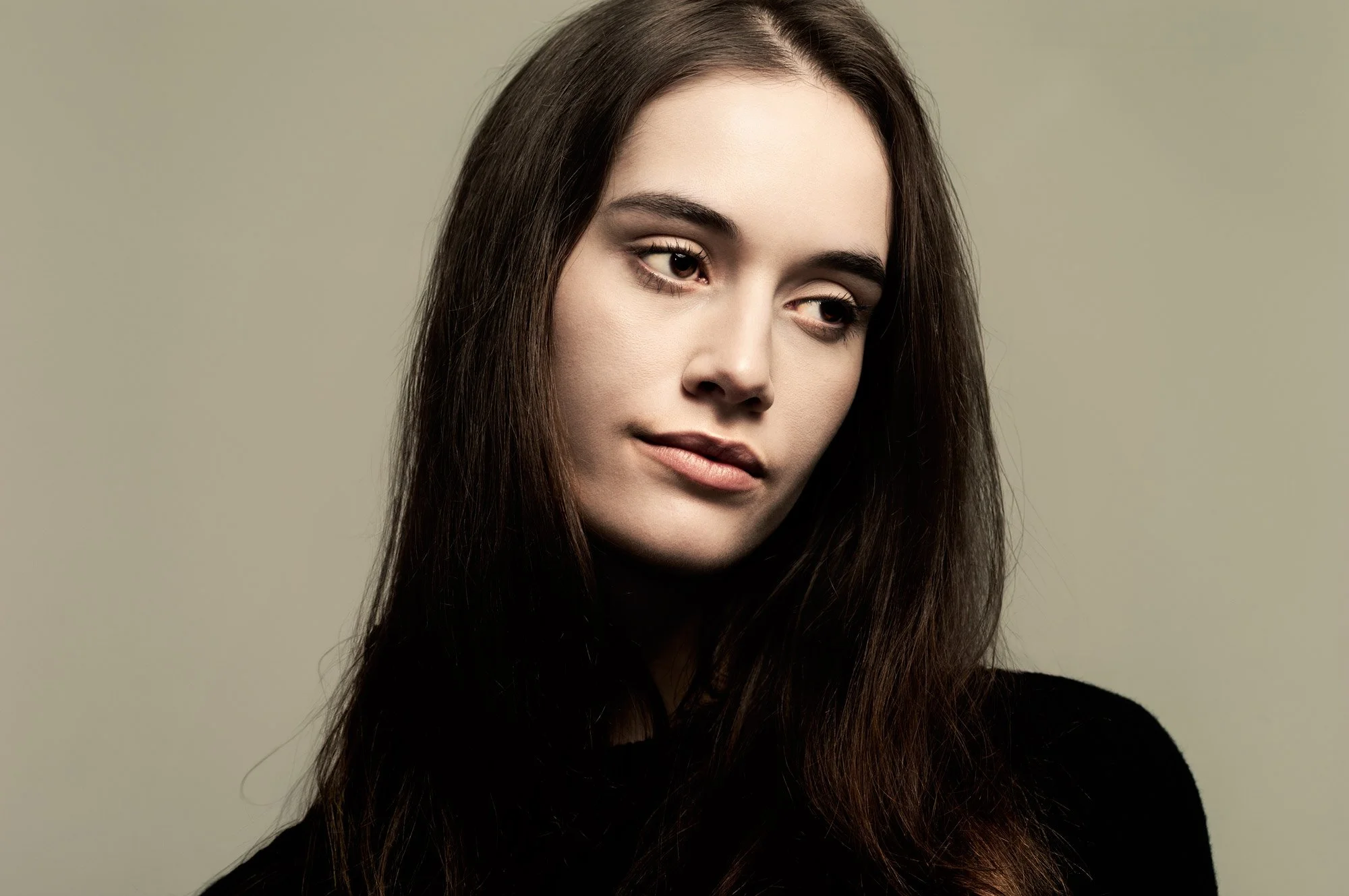 Close-up portrait of a young woman with long brown hair and green eyes, wearing a black top, against a muted green background.