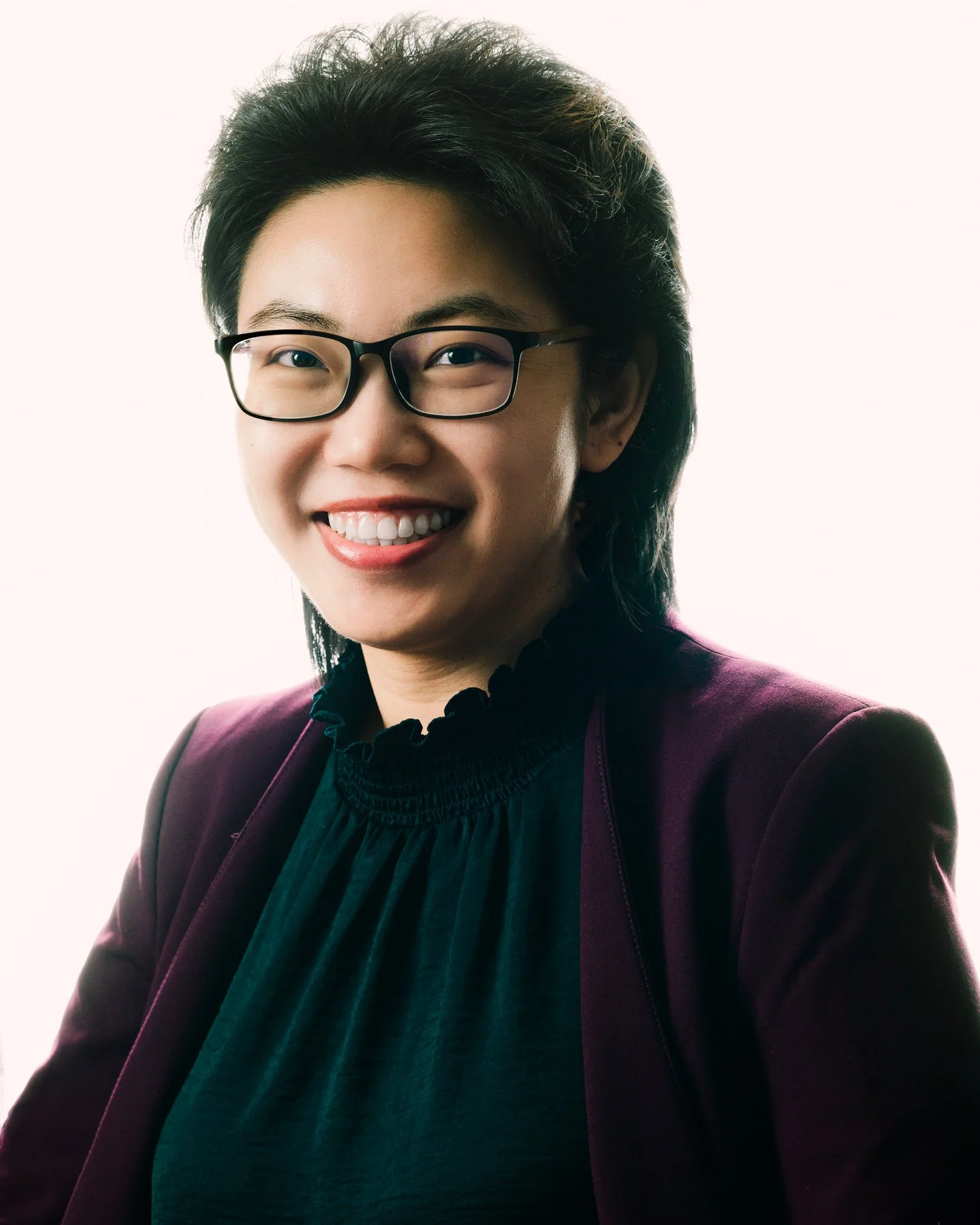 Smiling young Chinese symphony conductor, with eyewear and burgundy blazer, high-contrast lighting against neutral white background. Photographed at J Heroun’s Allston studio.