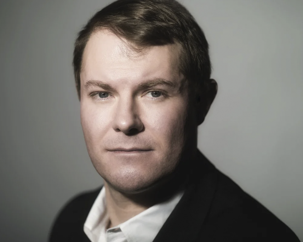 A man with light skin, short brown hair, blue eyes, dressed in a white shirt and black blazer, looking confidently at the camera against a plain gray background.