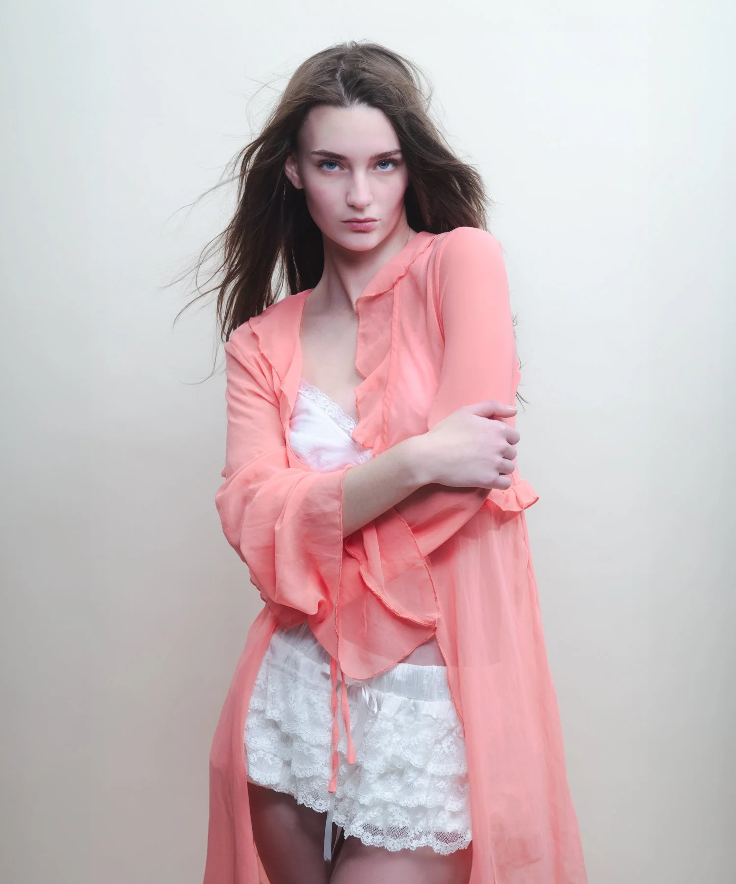 A young woman with long brown hair and blue eyes wearing a sheer pink robe over white lace lingerie standing against a plain off-white background.