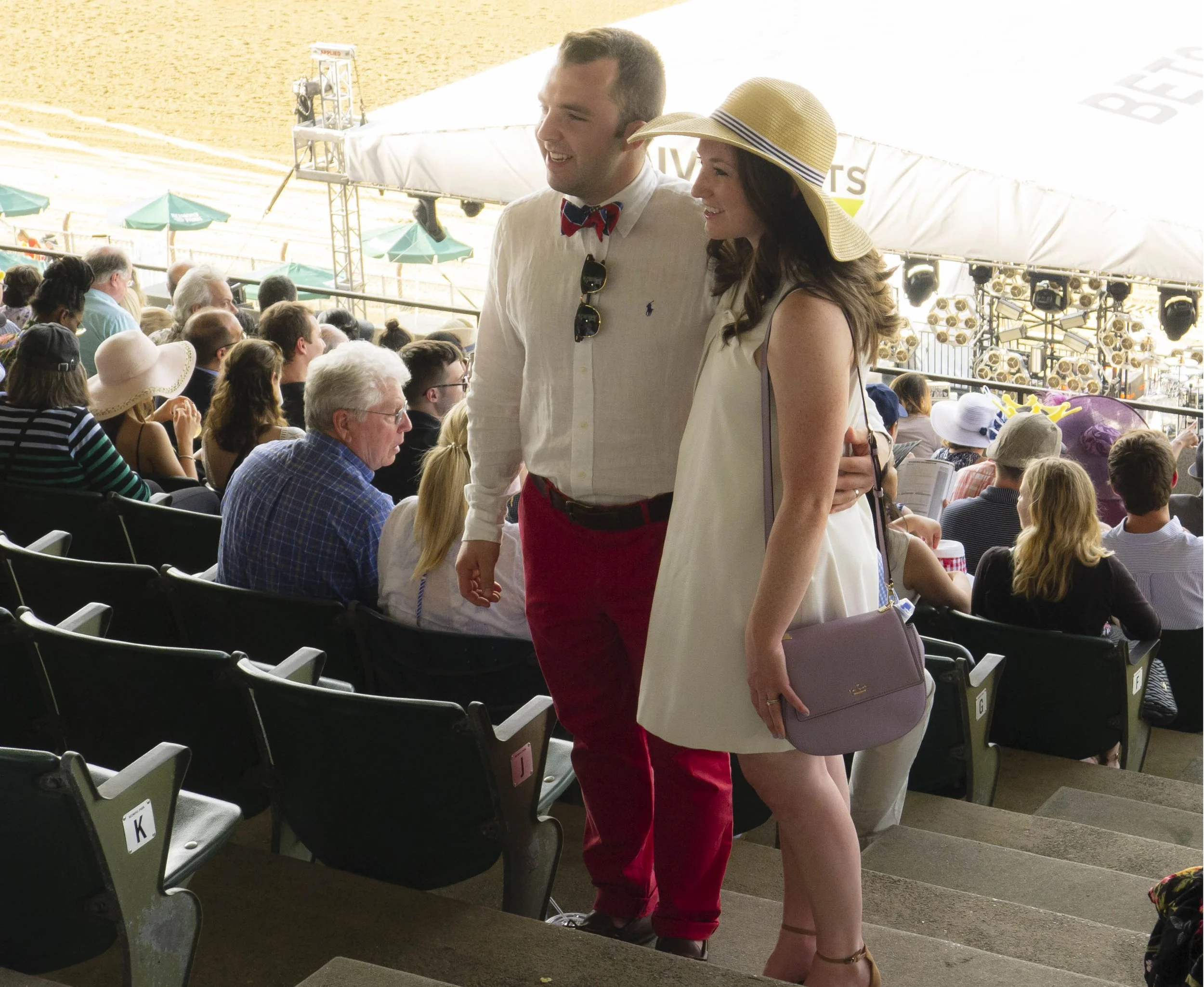 Couple at a crowded outdoor event, standing and smiling, with the man wearing a white shirt and red pants, and the woman in a cream dress and wide-brimmed yellow hat, surrounded by stadium seating and many other attendees.