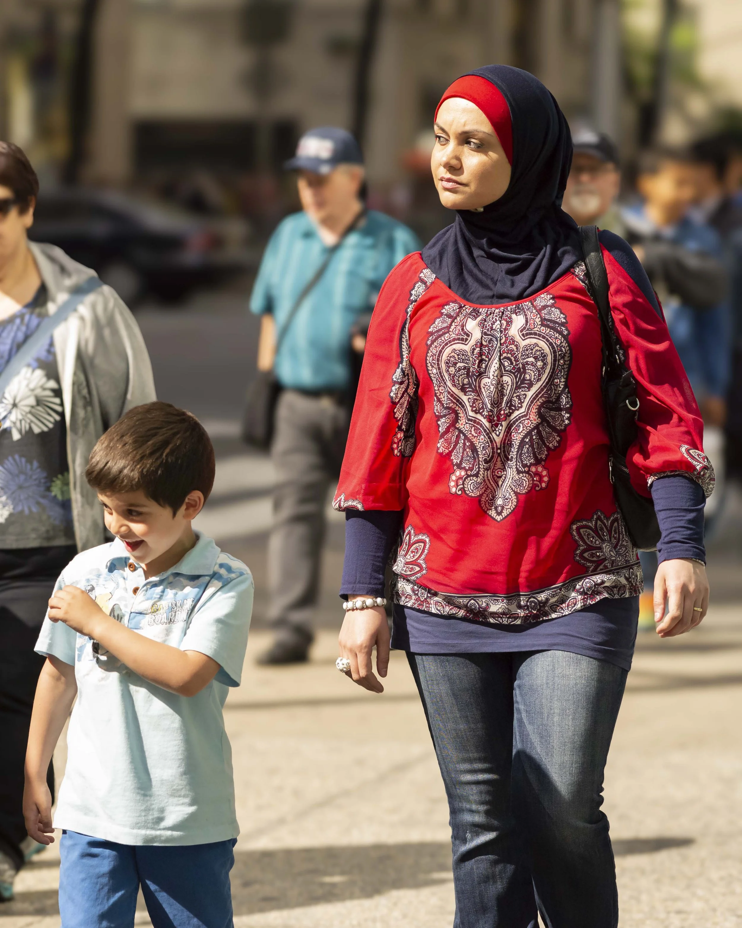 A woman wearing a black hijab with a red cap underneath, a red jacket with paisley patterns, and blue jeans walking outdoors with children and other people in the background.