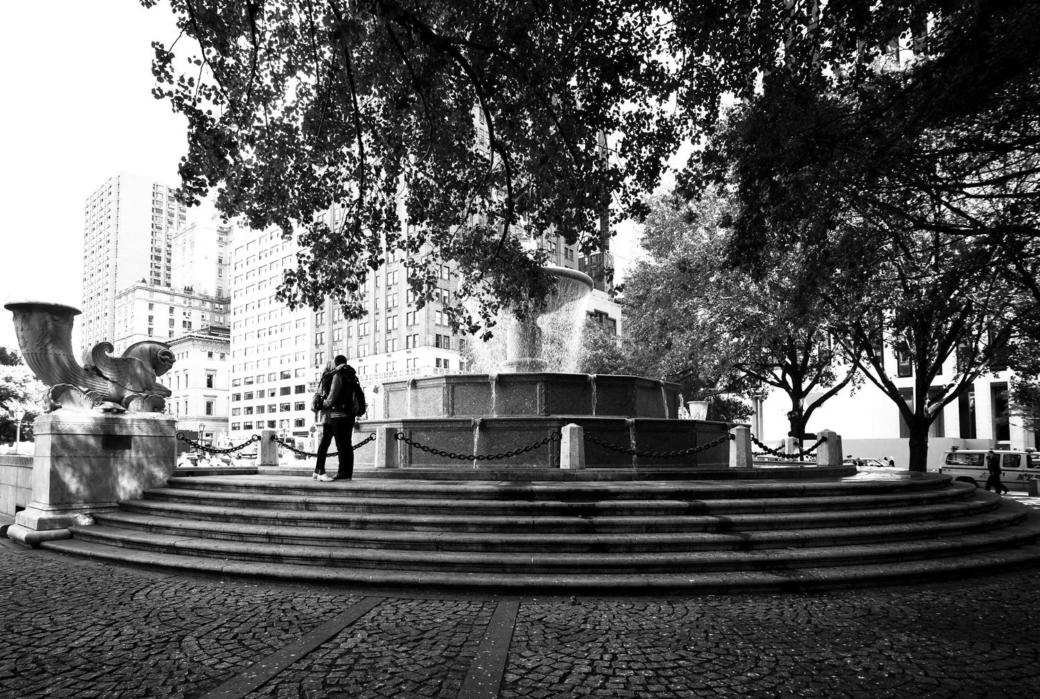 Black and white image of a city park with a decorative fountain surrounded by trees, with skyscrapers in the background. Two people stand near the fountain, under the trees.