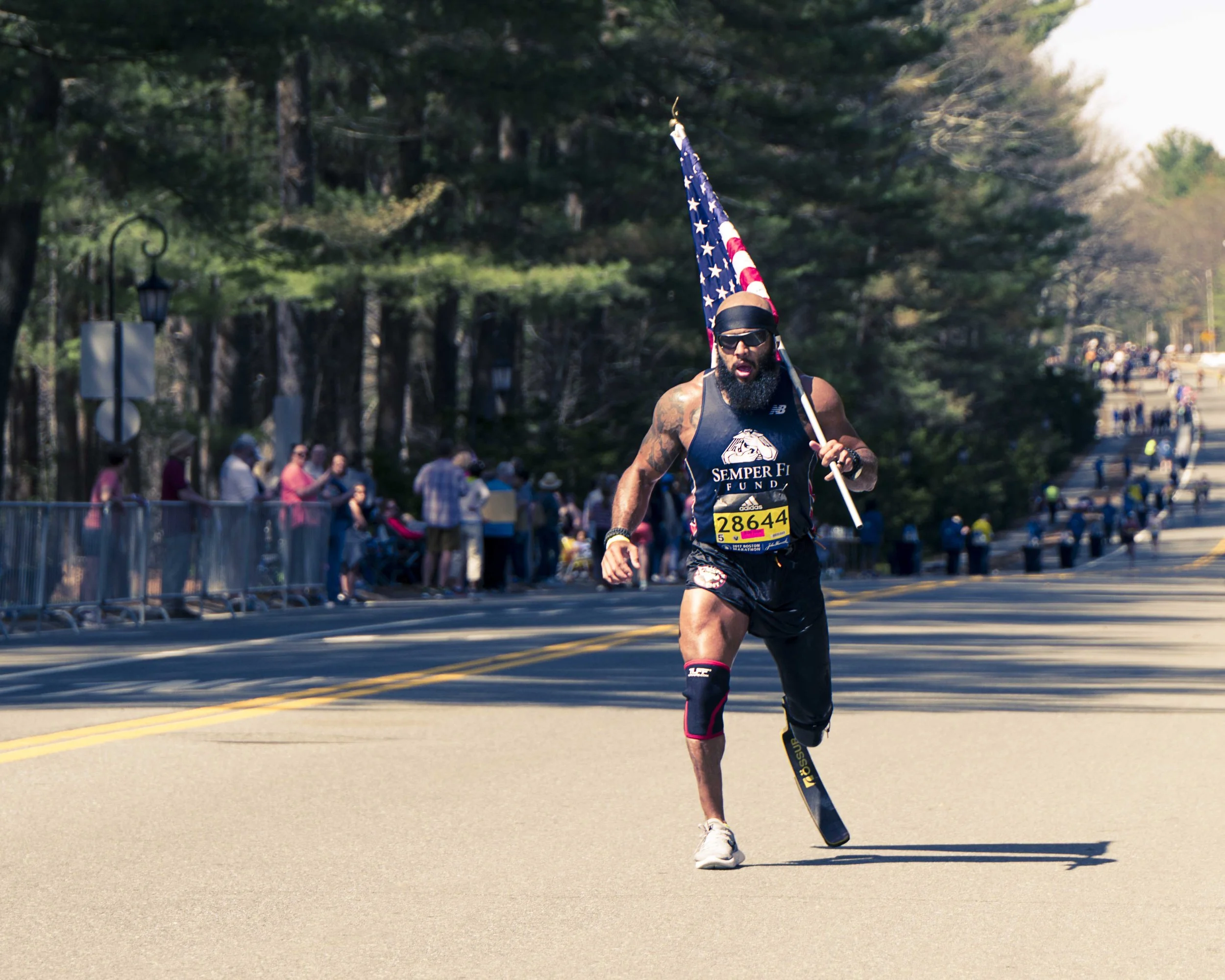 A man running and holding an American flag during a marathon or race in a park, with spectators watching along the side.